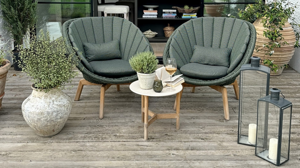Two shell-shaped outdoor lounge chair in dark green with with marble look-a-like side table with teak base, outdoor accessories of two black lanterns and plants in ceramic planters, terrace decor 