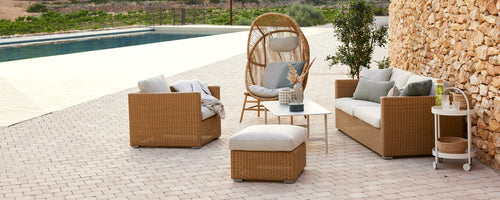 Stylish outdoor seating area featuring woven sofas, a hanging chair, and a footstool with cushions, surrounded by greenery.
