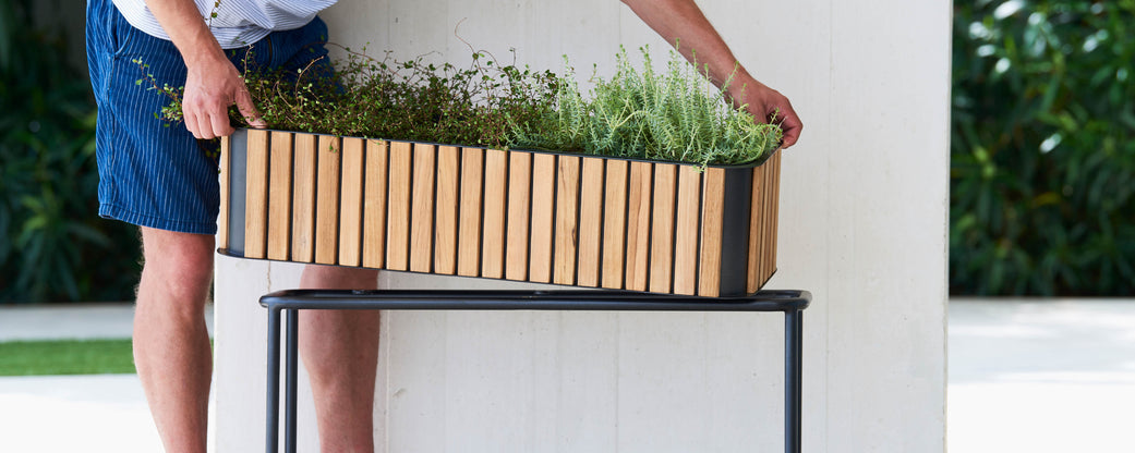 Person adjusting a planter box filled with grass and plants, positioned on a stand. The planter features a wooden slatted design.