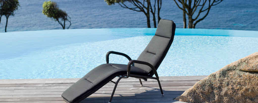 A sleek lounge chair by a tranquil pool, with a stunning view of the water and gentle trees against a vibrant sunrise backdrop.