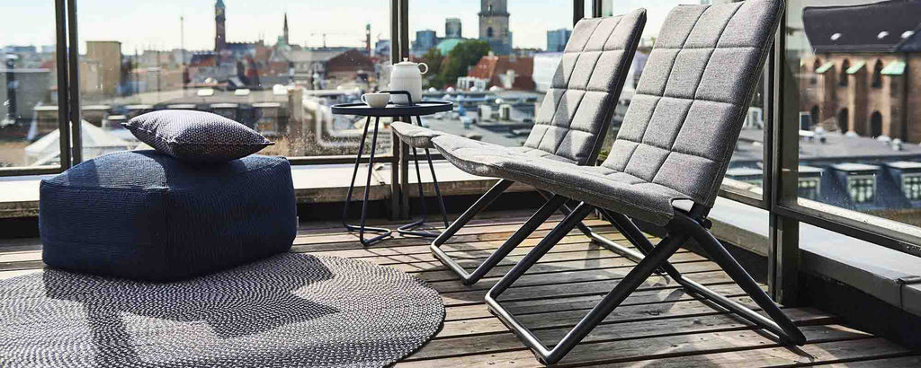 A modern folding lounge chair paired with a pillow and a round rug, set against a city skyline backdrop.