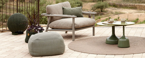 Stylish seating area featuring a comfortable chair, round table, pouf, and decorative accessories arranged on a textured rug.