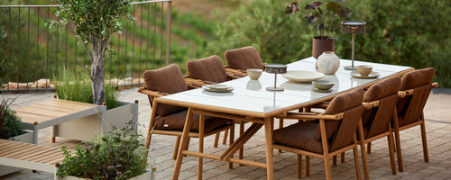 A stylish dining table set with brown chairs, elegant tableware, and decorative elements, surrounded by greenery.