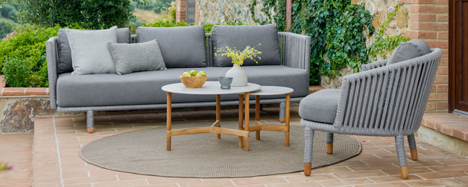 Stylish gray outdoor sofas with cushions, accompanied by a round coffee table and a vase with flowers, set in a lush green environment.