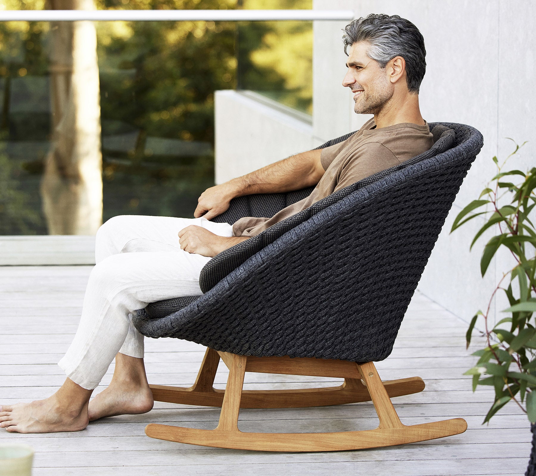 A man sits relaxed in a stylish, modern rocking chair, featuring a woven design and a natural wood base, surrounded by greenery.