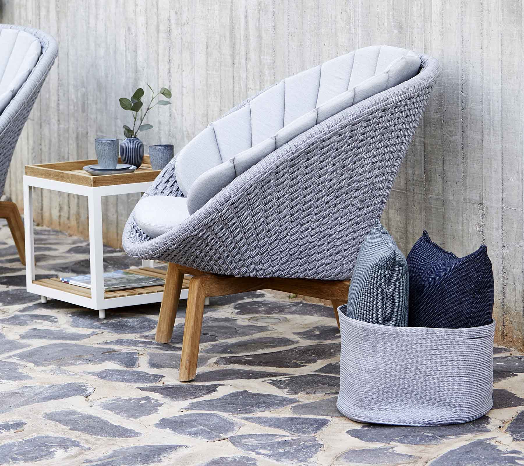 Modern grey lounge chair with a woven design, accompanied by pillows and a small wooden side table. Stone floor enhances the aesthetic.