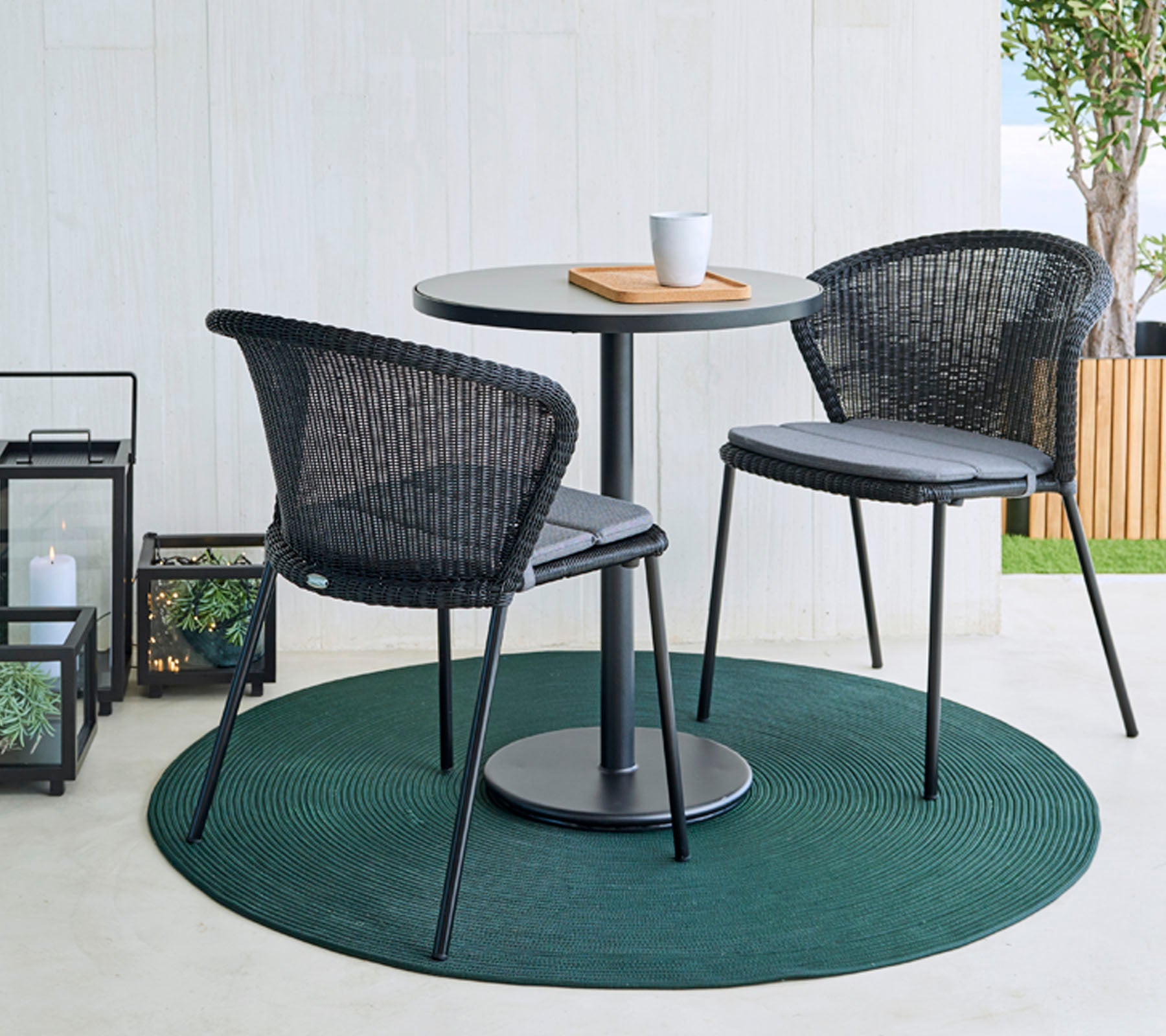 A round table with two black woven chairs on a green rug, accompanied by a coffee cup and a wooden board. Decorative lanterns in the background.