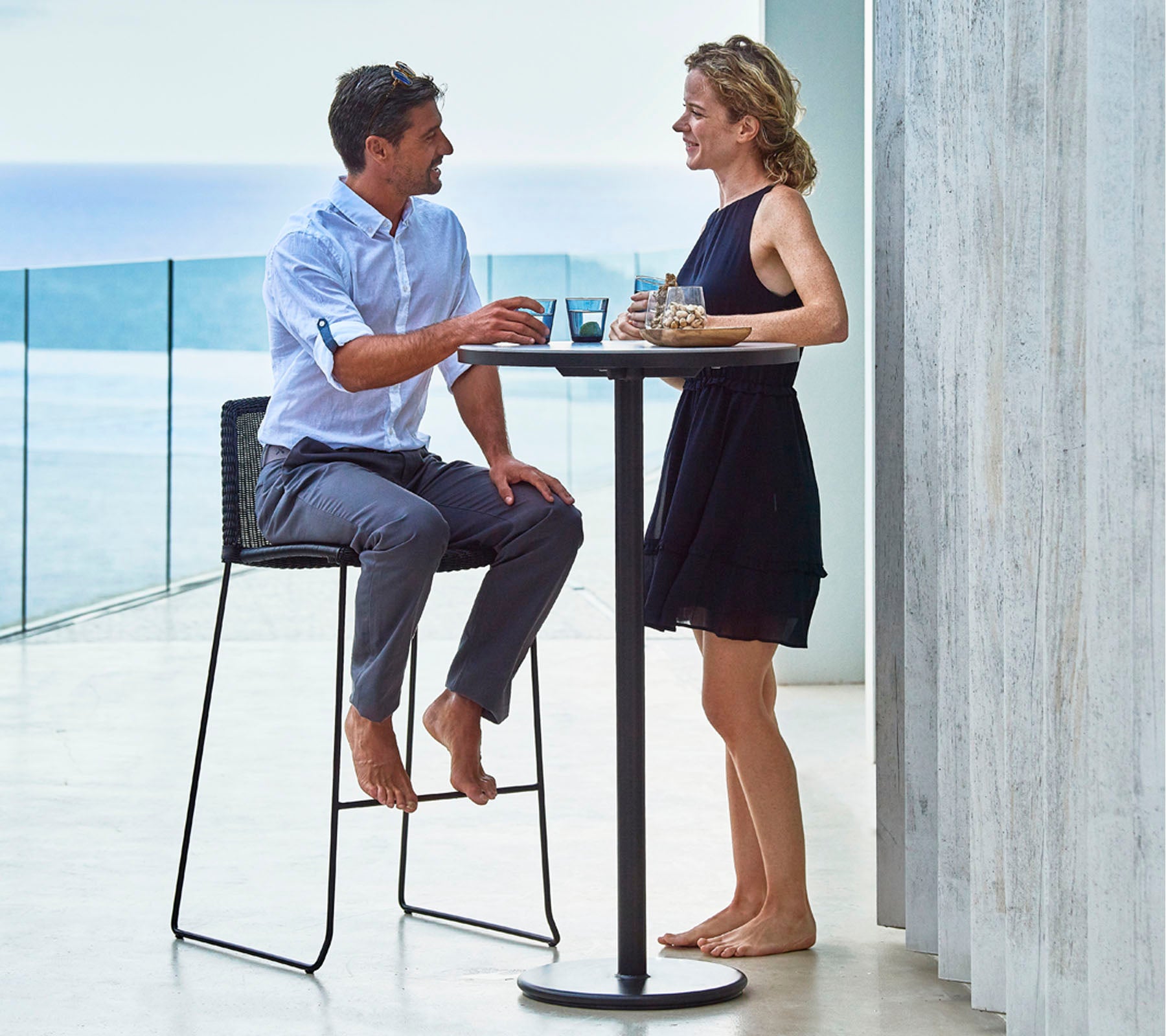 A man and woman enjoying drinks at a tall table, engaged in conversation, with a scenic backdrop of water in the distance.