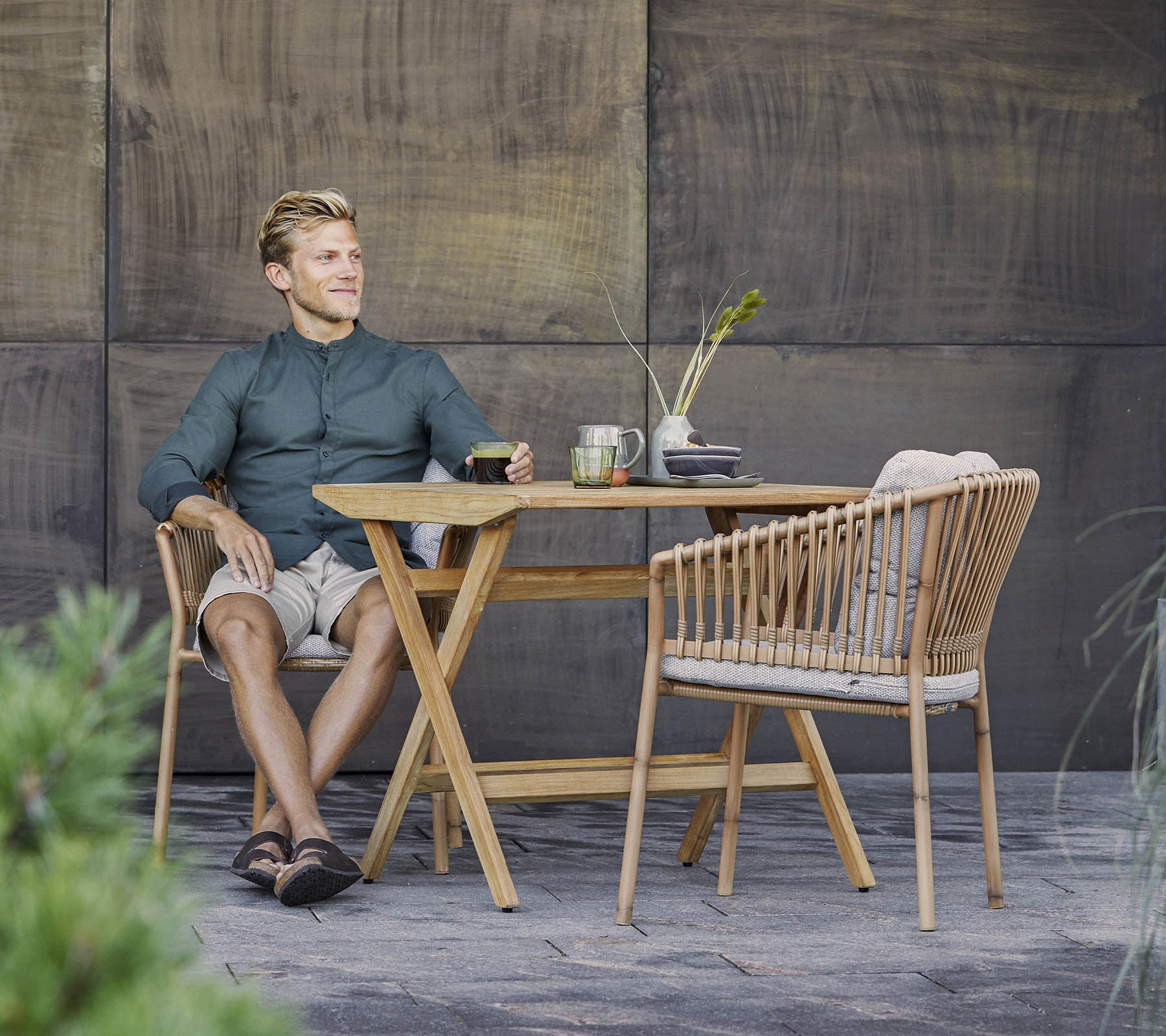 A man sits relaxed at a stylish wooden table with a light drink, surrounded by greenery and a textured backdrop.