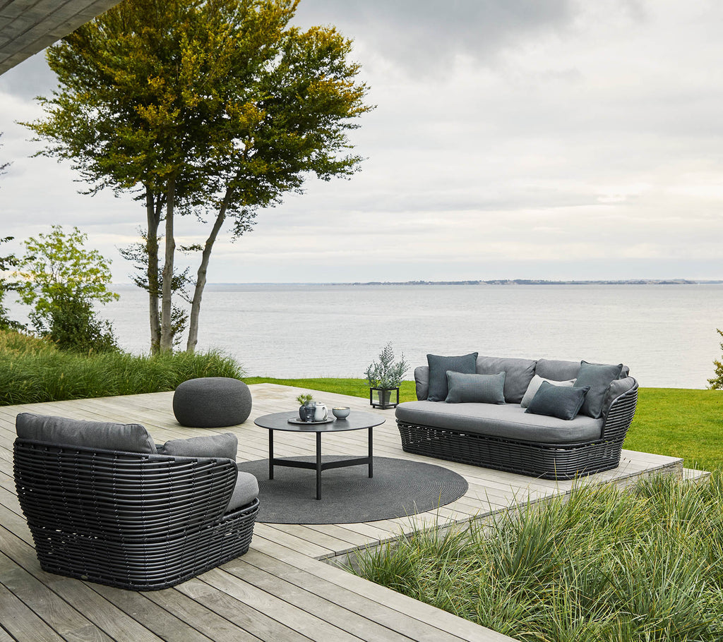 Cozy seating arrangement with a sofa and chairs set around a circular table, surrounded by greenery and a water view.