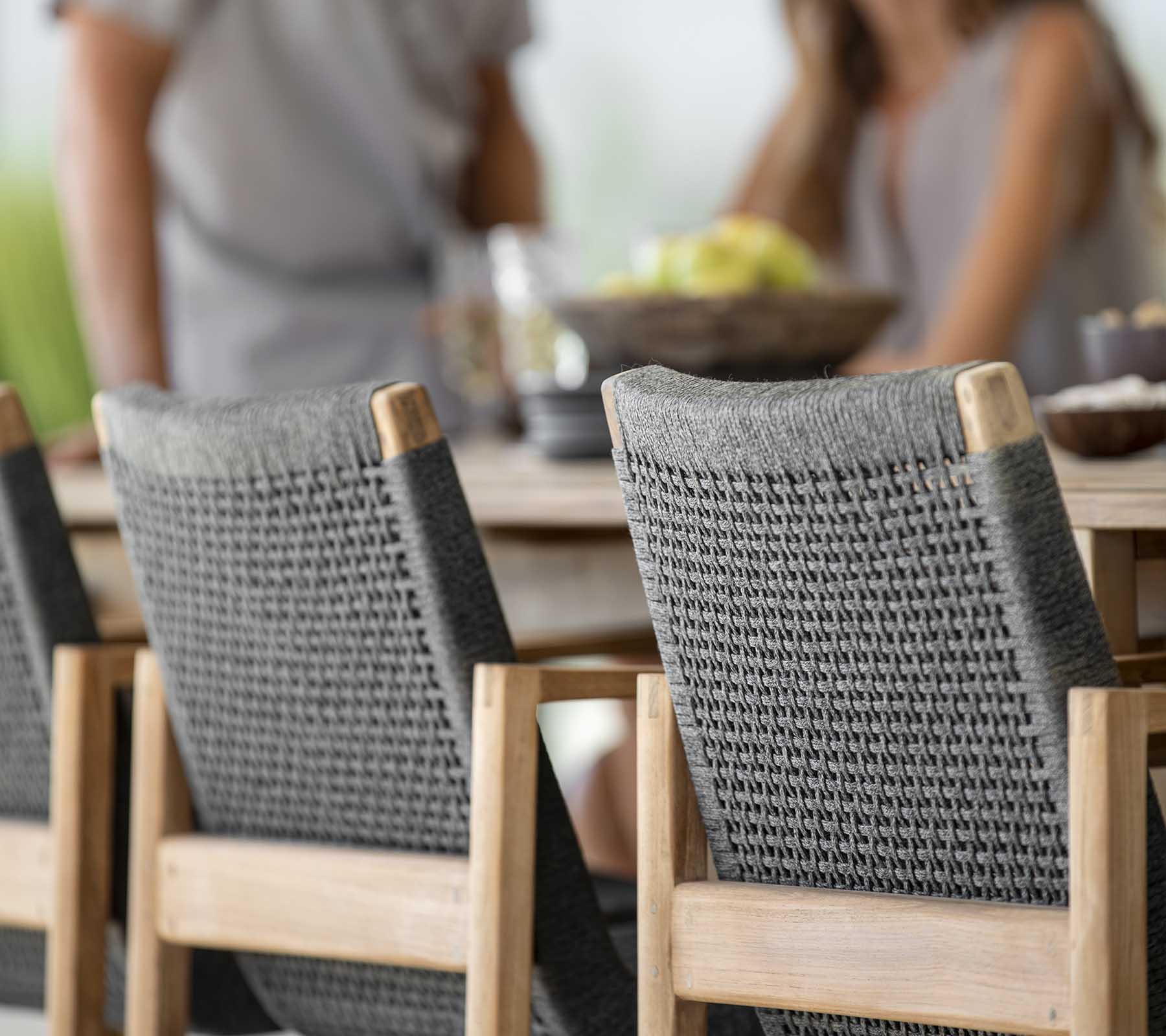Outdoor dining scene featuring a wooden table with two stylish gray woven chairs and people enjoying a meal together.