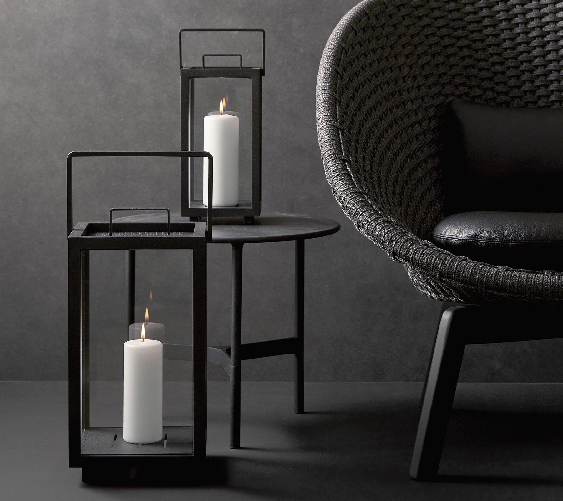 Two stylish candle lanterns with white candles, accompanied by a modern, textured chair on a minimalist table against a dark background.
