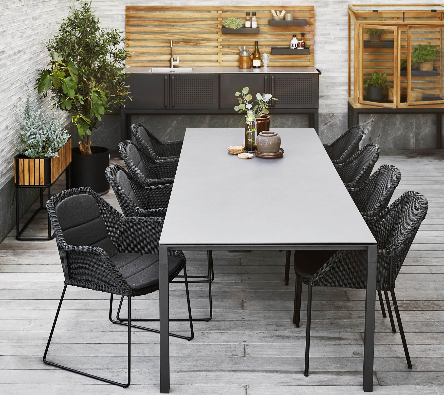 A modern dining setup featuring a long table surrounded by sleek black chairs, complemented by decorative plants and a wooden backdrop.