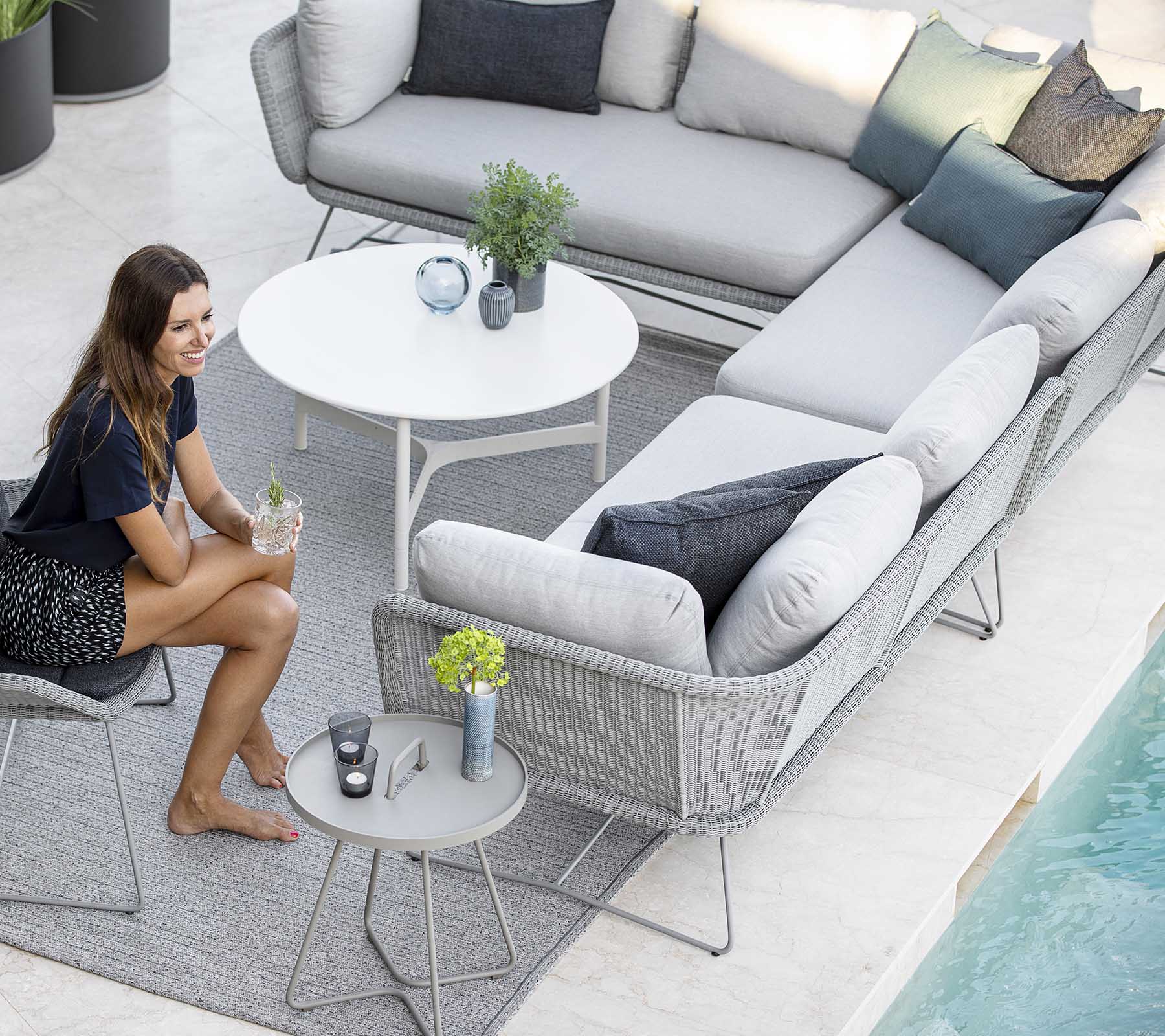 A stylish outdoor seating arrangement featuring a sofa, accent pillows, and a small table, complemented by plants and a pool nearby.