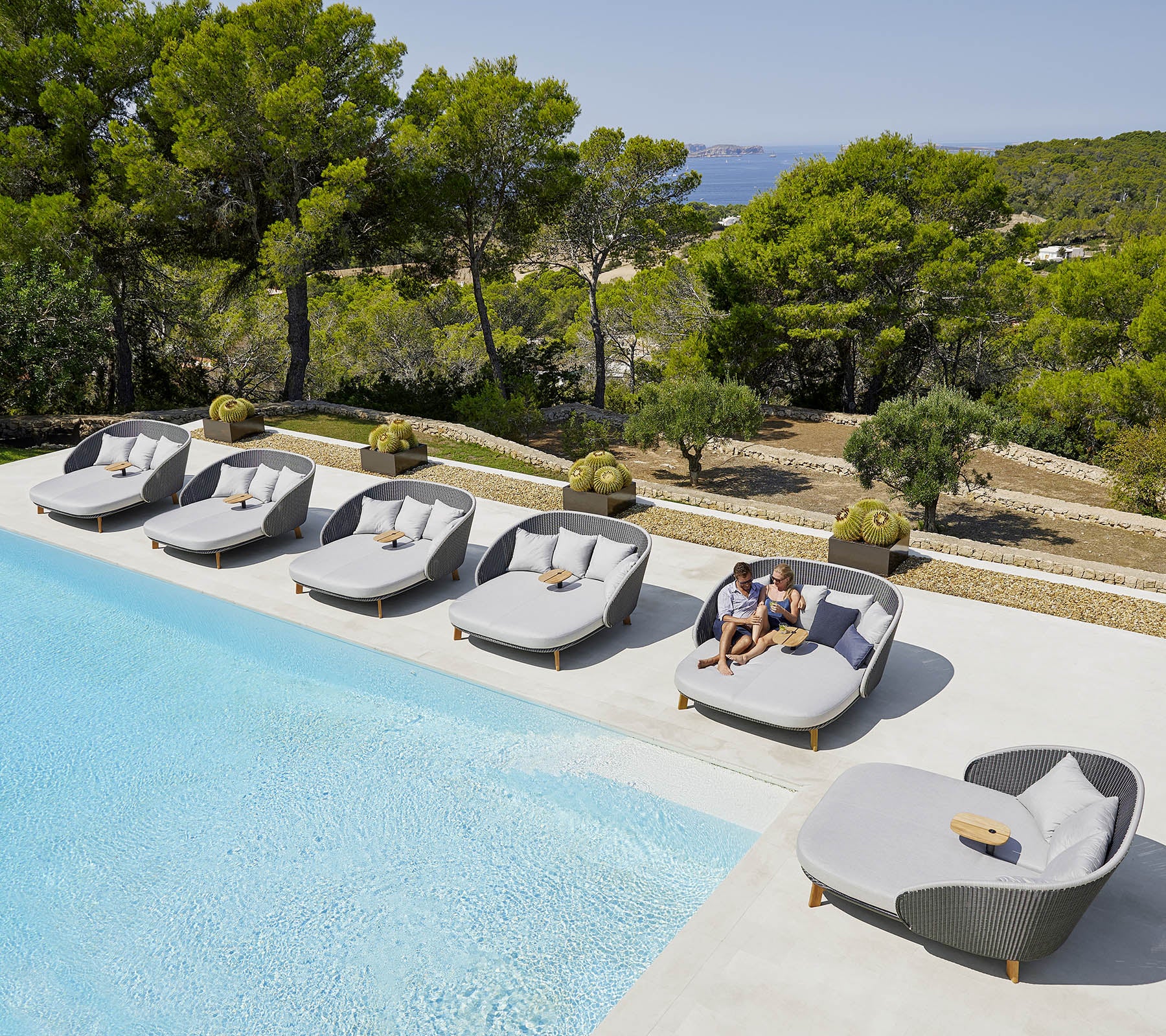 Lounge chairs arranged around a swimming pool with a scenic view of greenery and trees in the background. A person relaxes on one of the chairs.