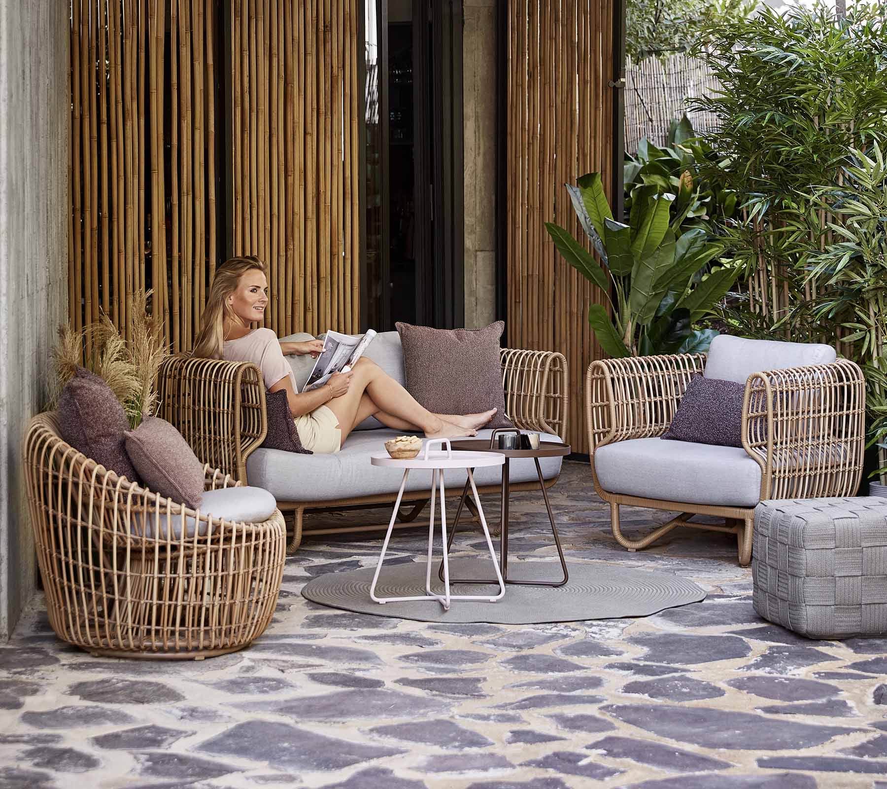 A stylish seating arrangement featuring woven furniture, cushions, and a coffee table, set among greenery and decorative elements.