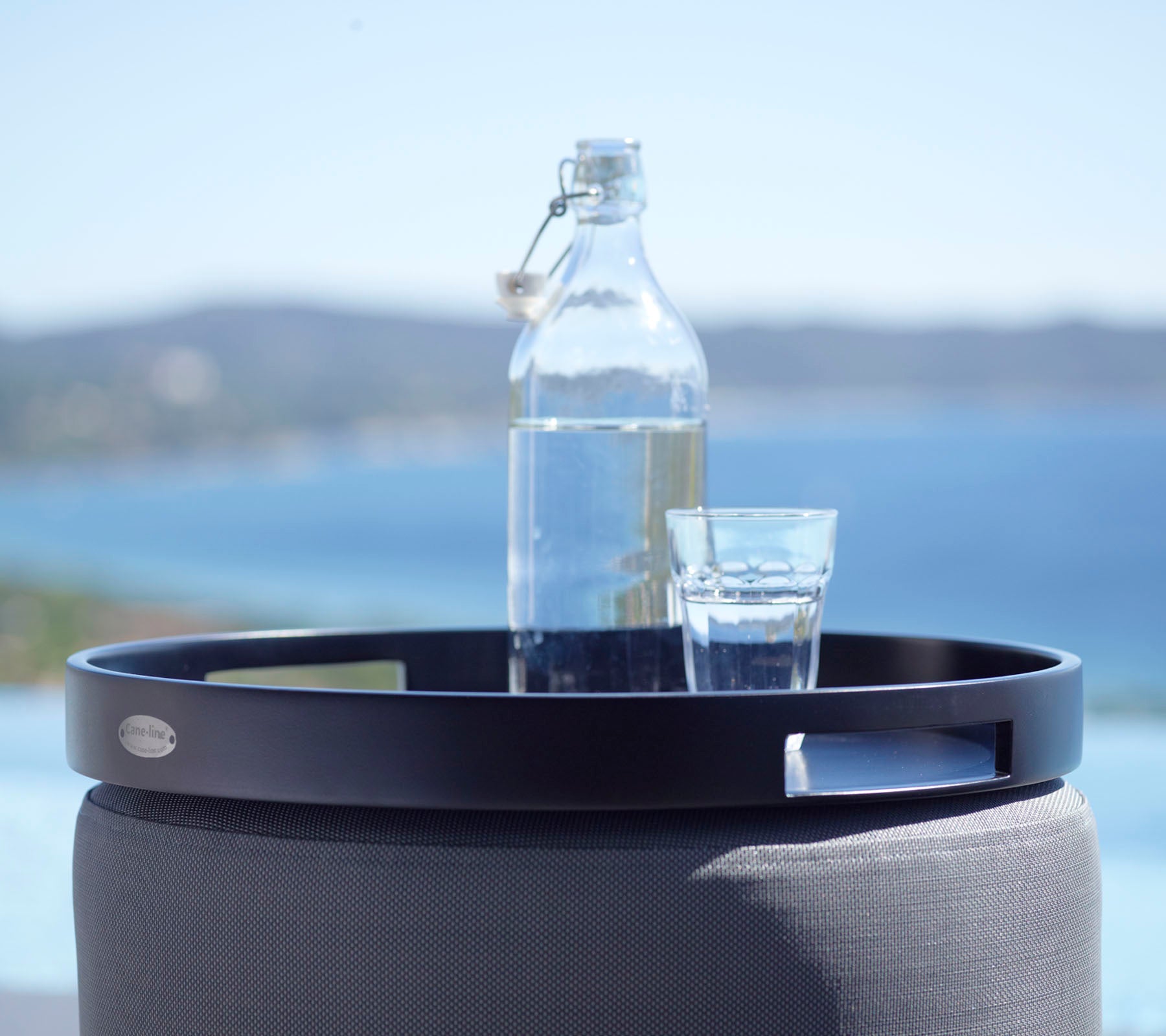 A black round tray holds a glass of water and a glass bottle, set against a scenic backdrop of the sea and distant hills.