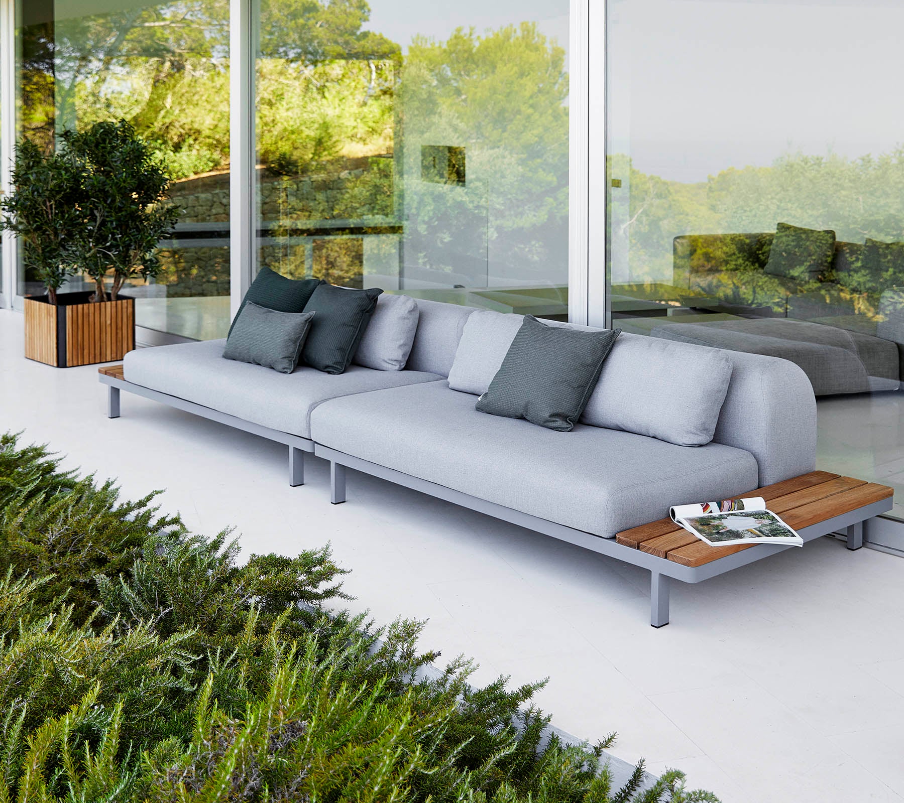 Modern sofa with light gray cushions and wooden accents, accompanied by decorative pillows and surrounded by greenery.