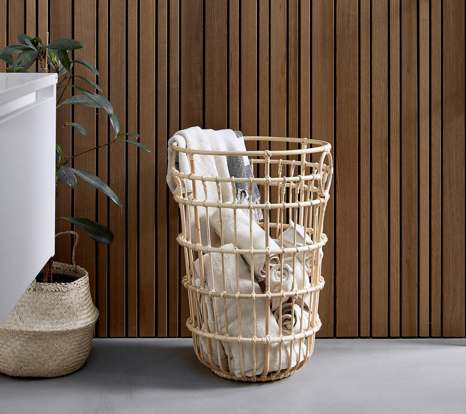 Woven laundry basket filled with rolled towels, placed beside a plant and a textured wooden wall.