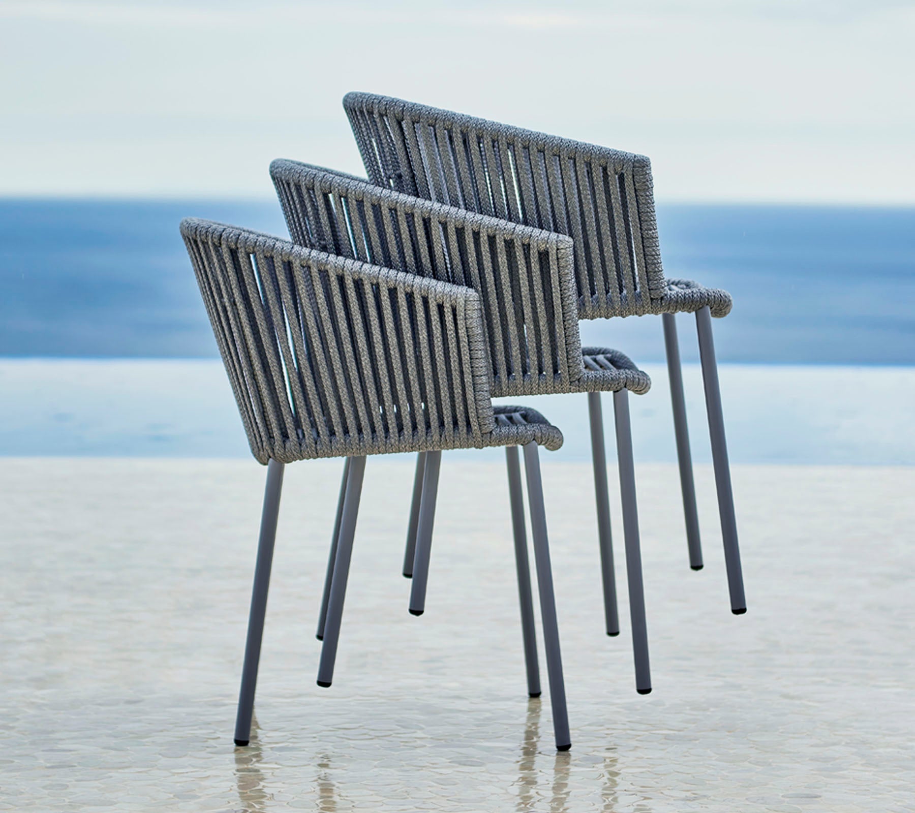 Three modern chairs with a stylish design, featuring a slatted back and armrests, positioned beside a serene water view.