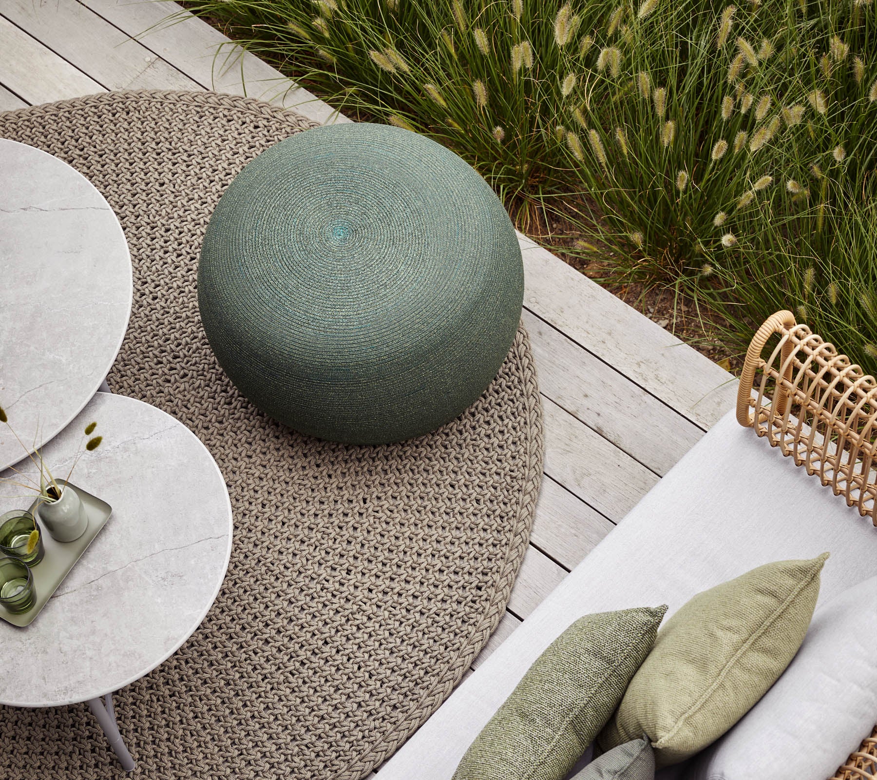 A cozy outdoor seating area featuring a round green pouf, circular tables, and textured cushions on a neutral rug. Lush greenery in the background.