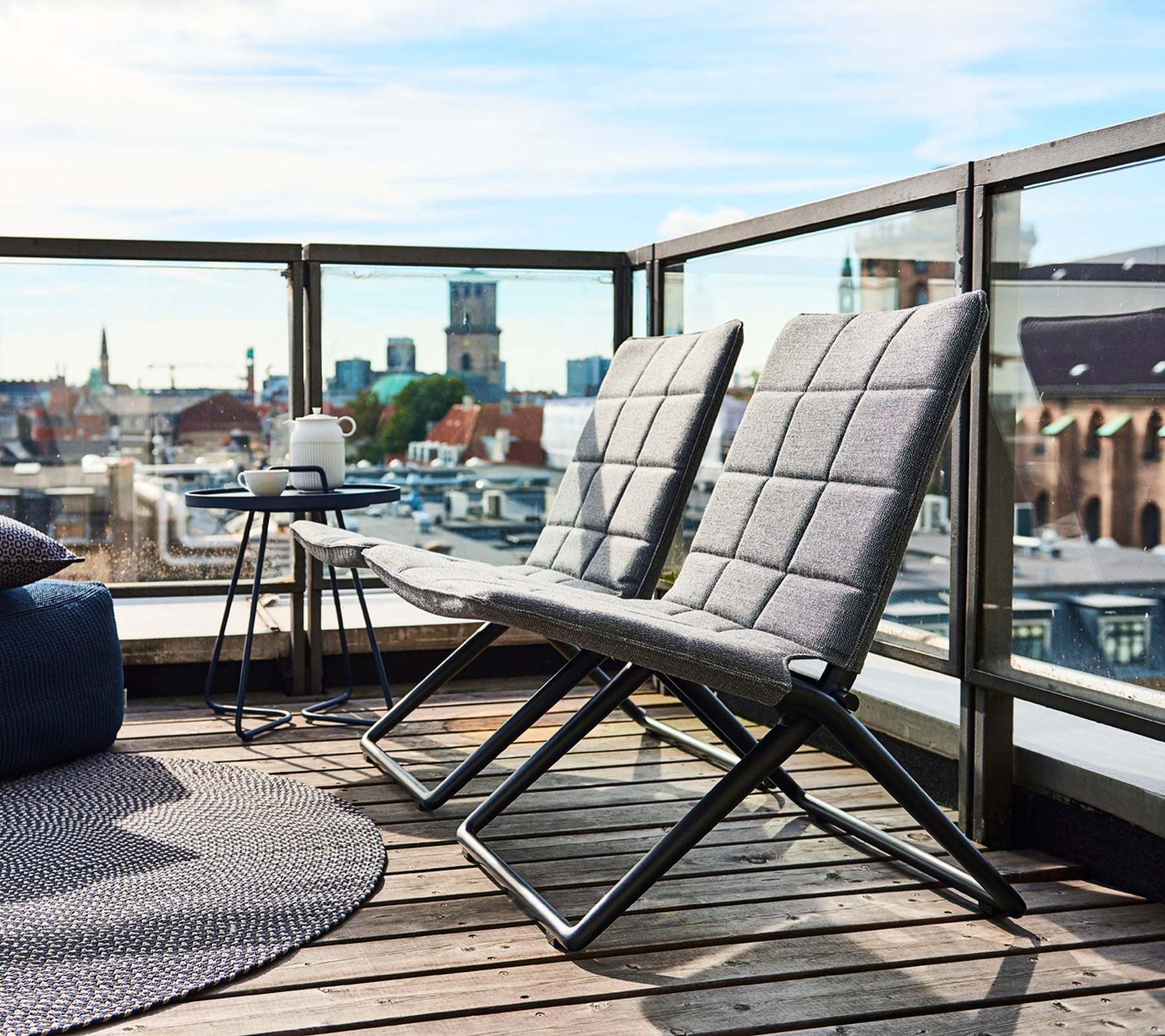 Two modern lounge chairs with sleek frames and textured cushions, accompanied by a small side table and cityscape views in the background.