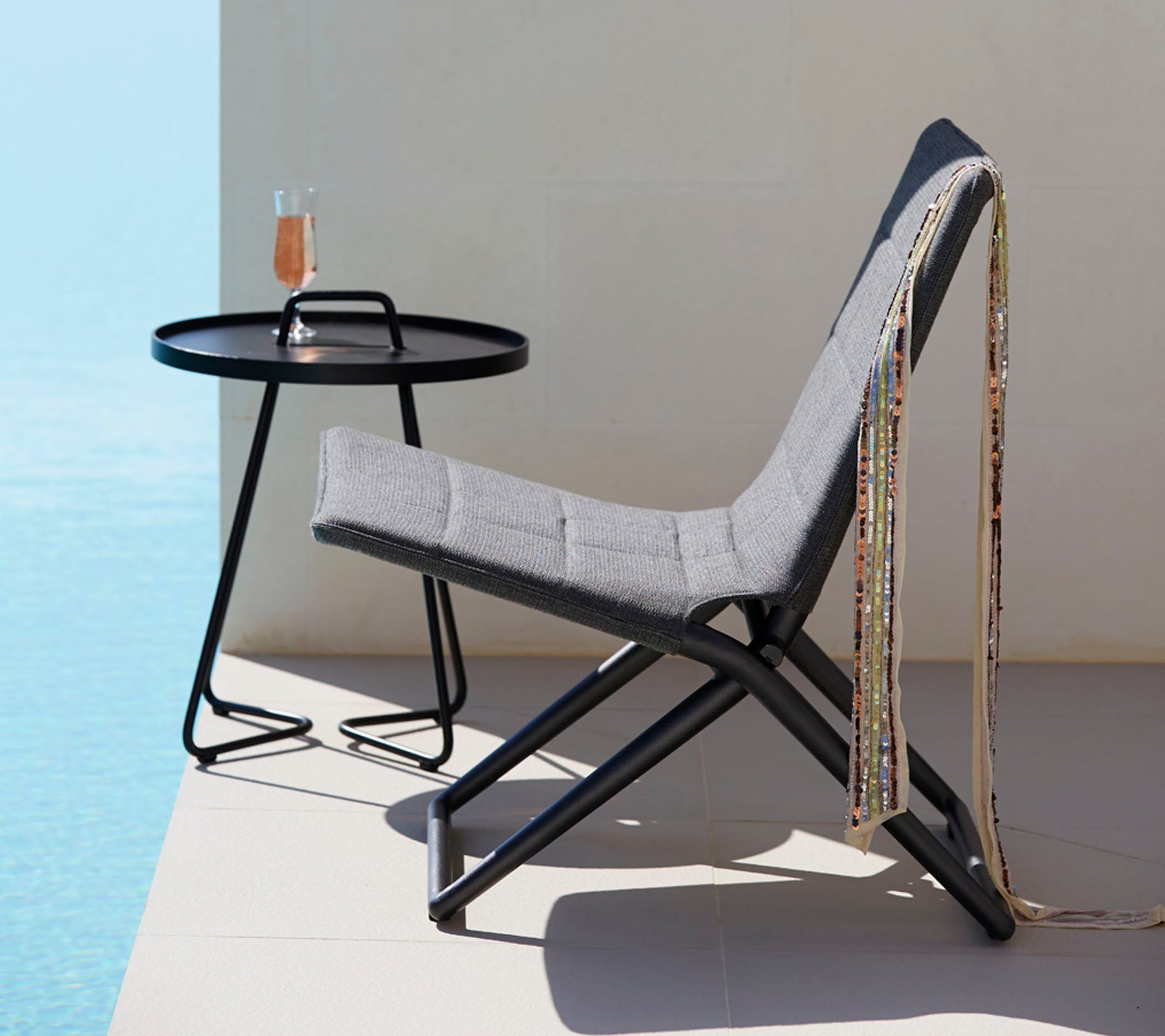 A stylish chair with gray fabric and a sleek black metal frame, paired with a round table holding a beverage and decorative beads.