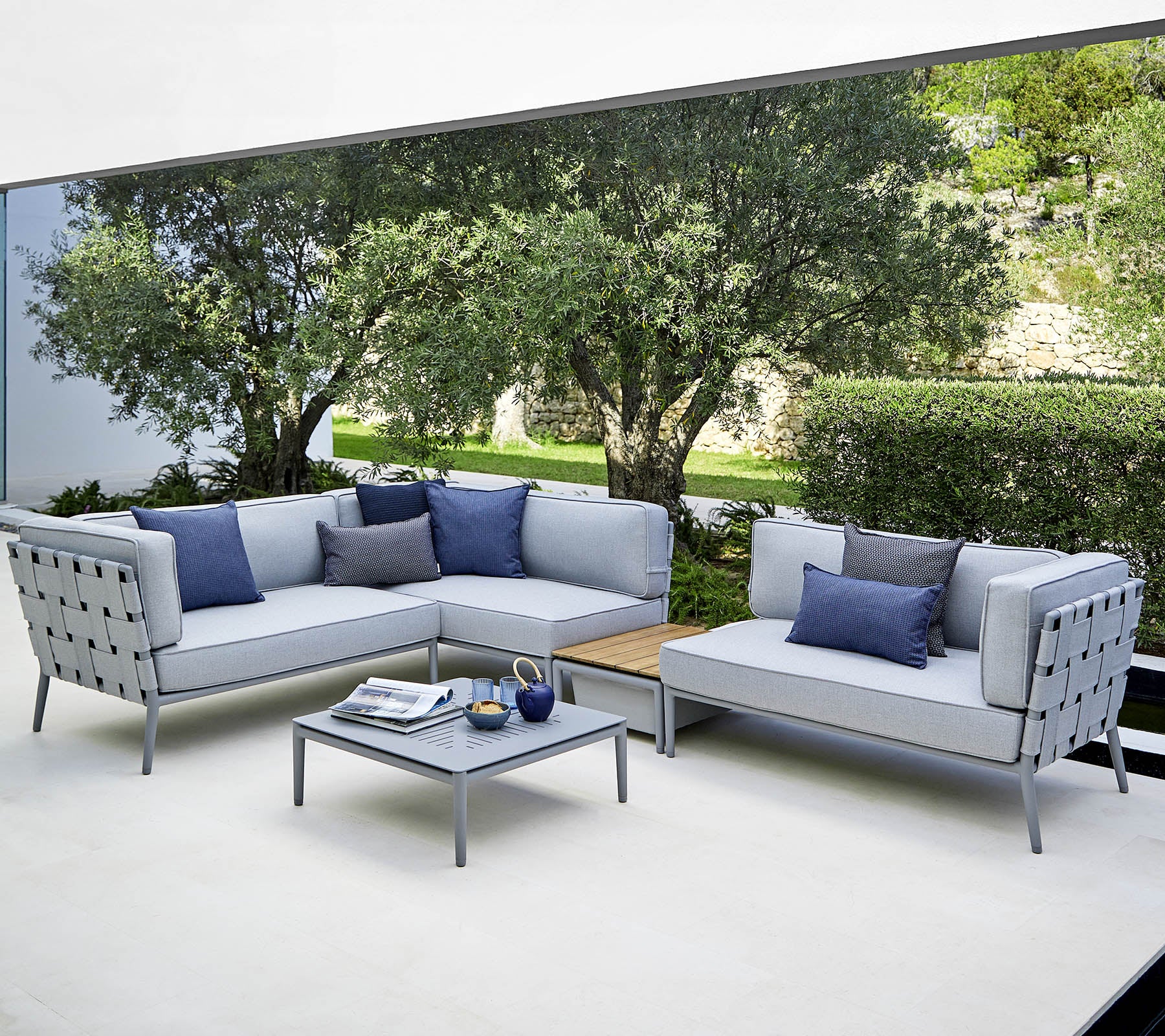 A modern outdoor seating arrangement featuring light gray sofas with blue cushions and a matching coffee table, set against a lush background.