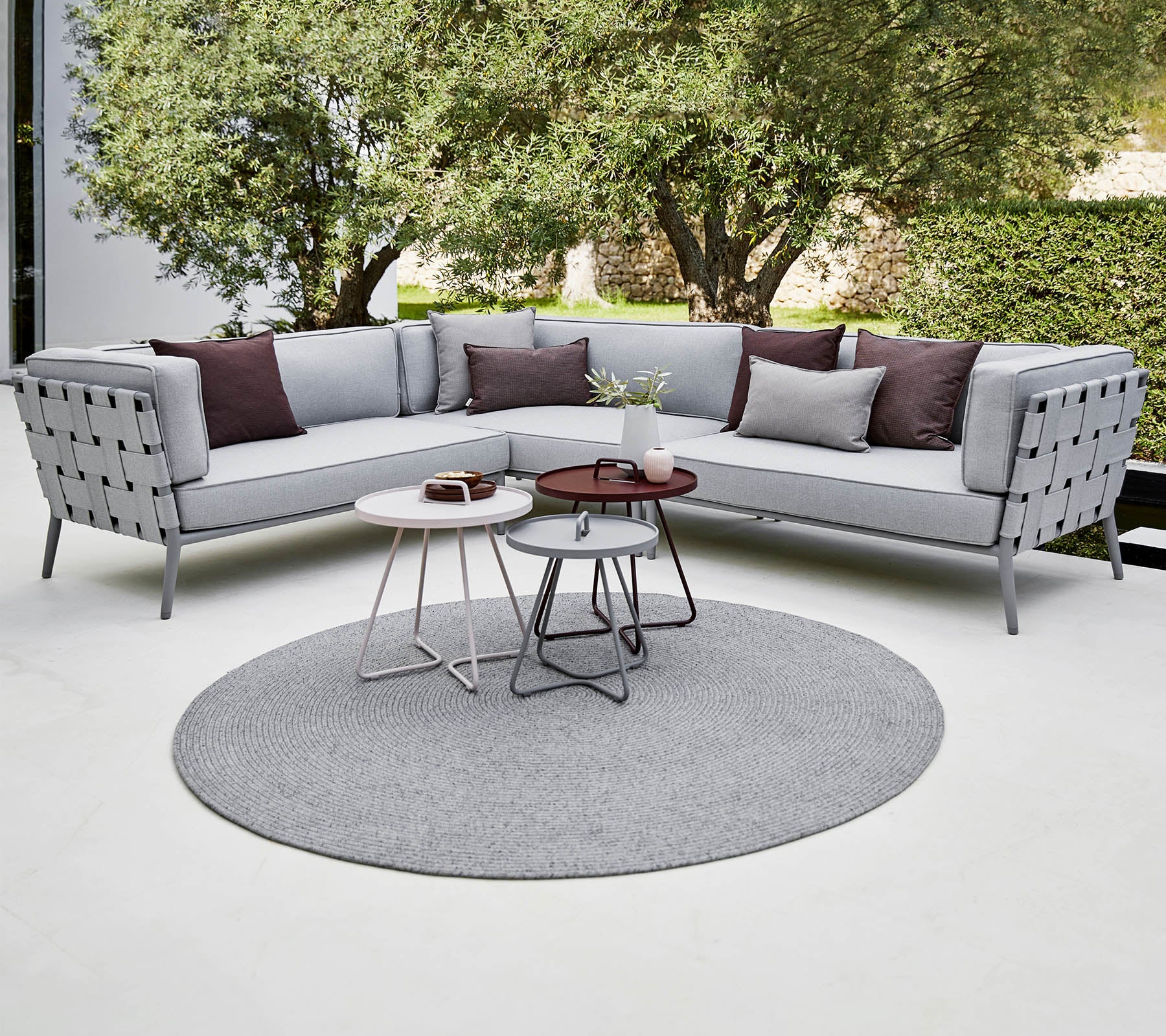 Modern sectional sofa in light gray with decorative pillows, accompanied by stylish coffee tables on a round rug. Lush greenery in the background.