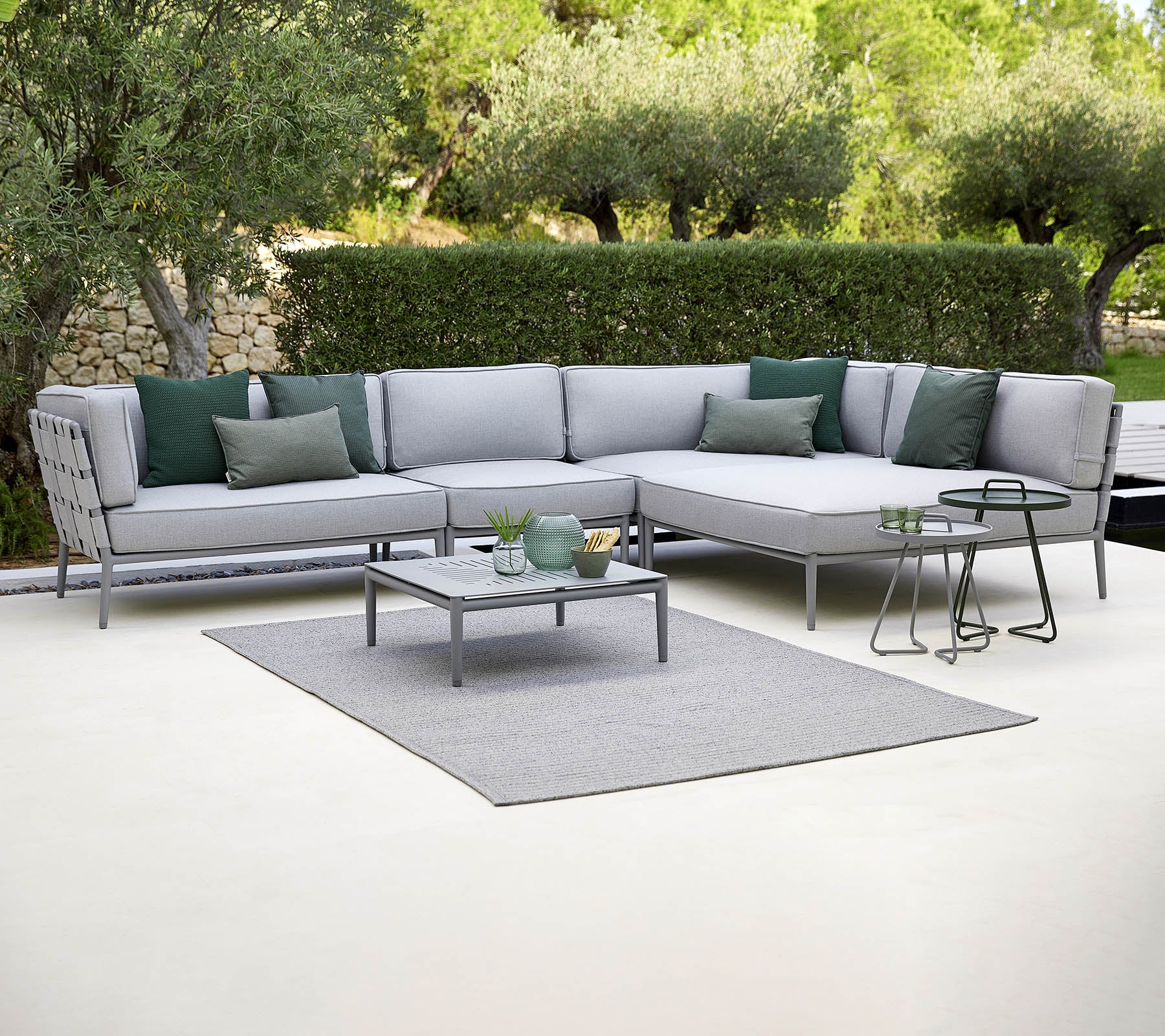Stylish outdoor sectional sofa with green cushions, a coffee table, and a rug, set against a backdrop of greenery.