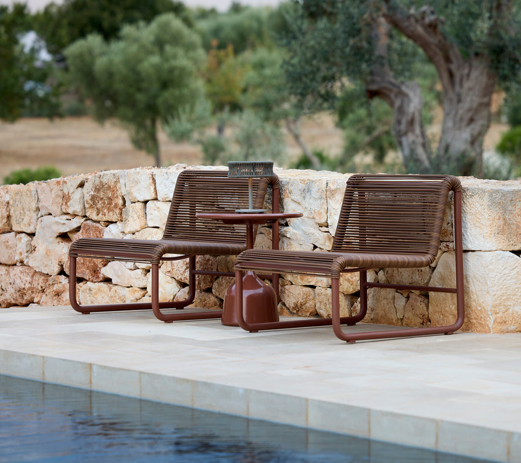 Two modern brown lounge chairs with a matching side table by a pool.