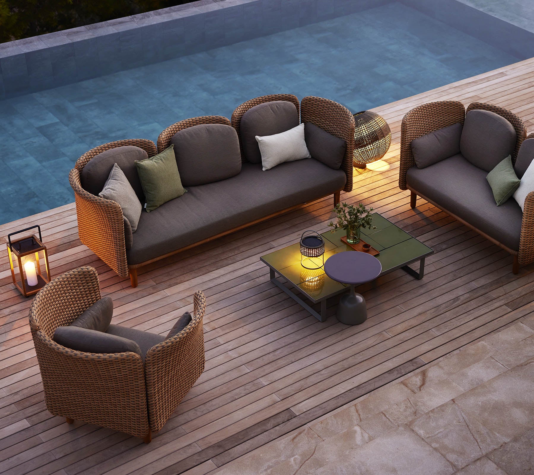 A cozy seating arrangement featuring a sofa and chairs with cushions, surrounded by greenery and a pool, on a wooden deck.