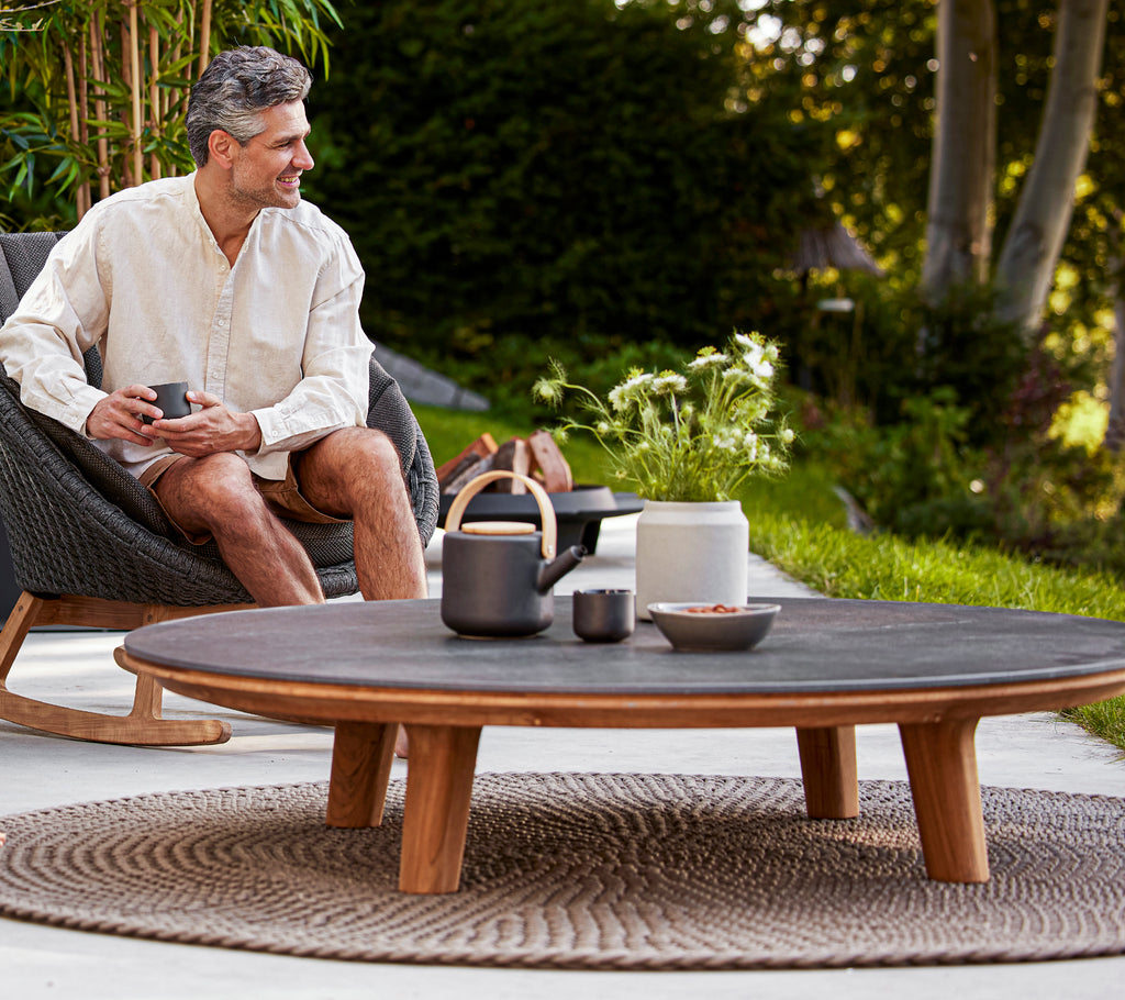 A man enjoys a drink while seated in a rocking chair beside a round coffee table adorned with stylish tableware and greenery.