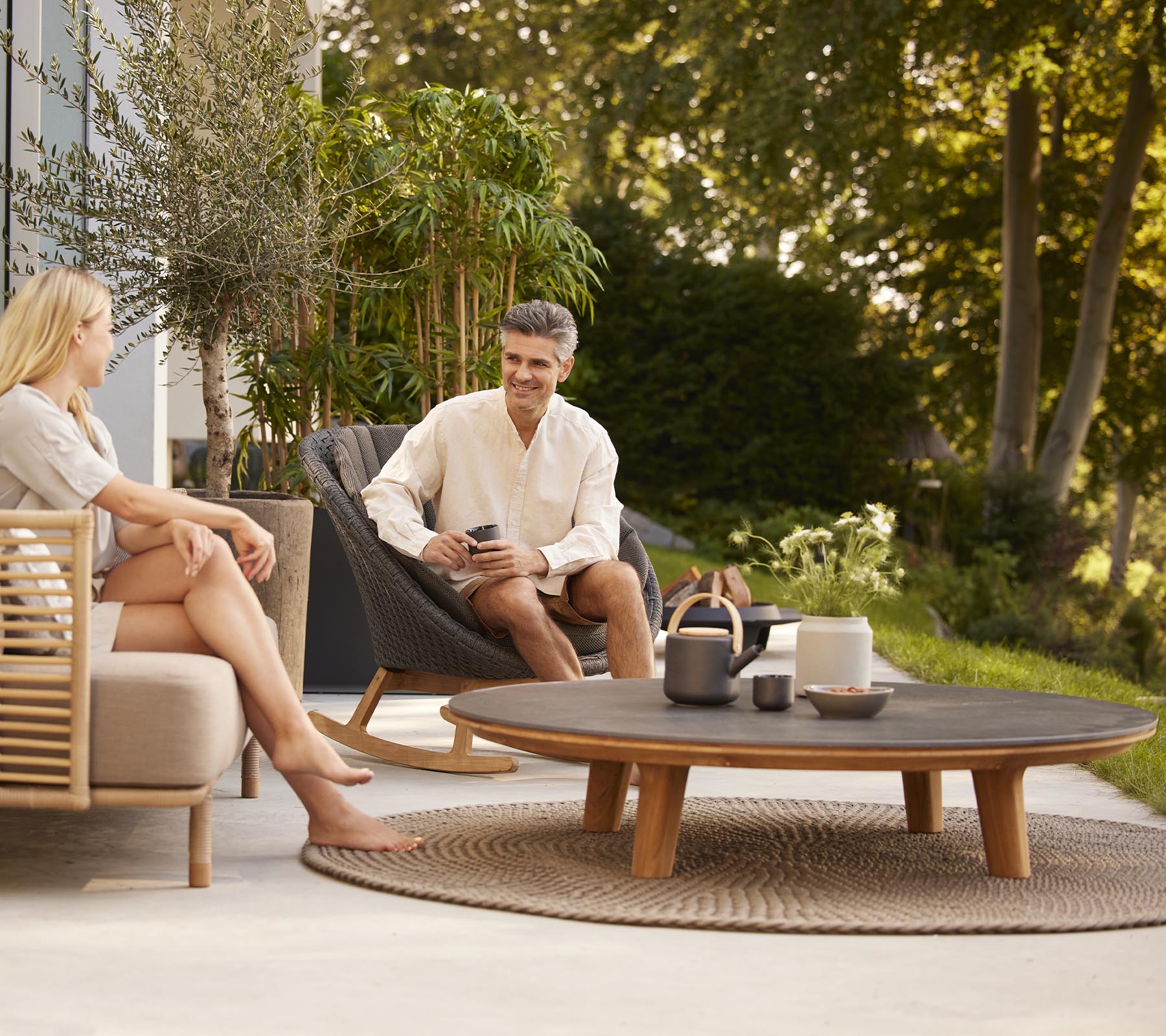 A man and woman relax on comfortable seating, enjoying drinks on a round coffee table surrounded by greenery.