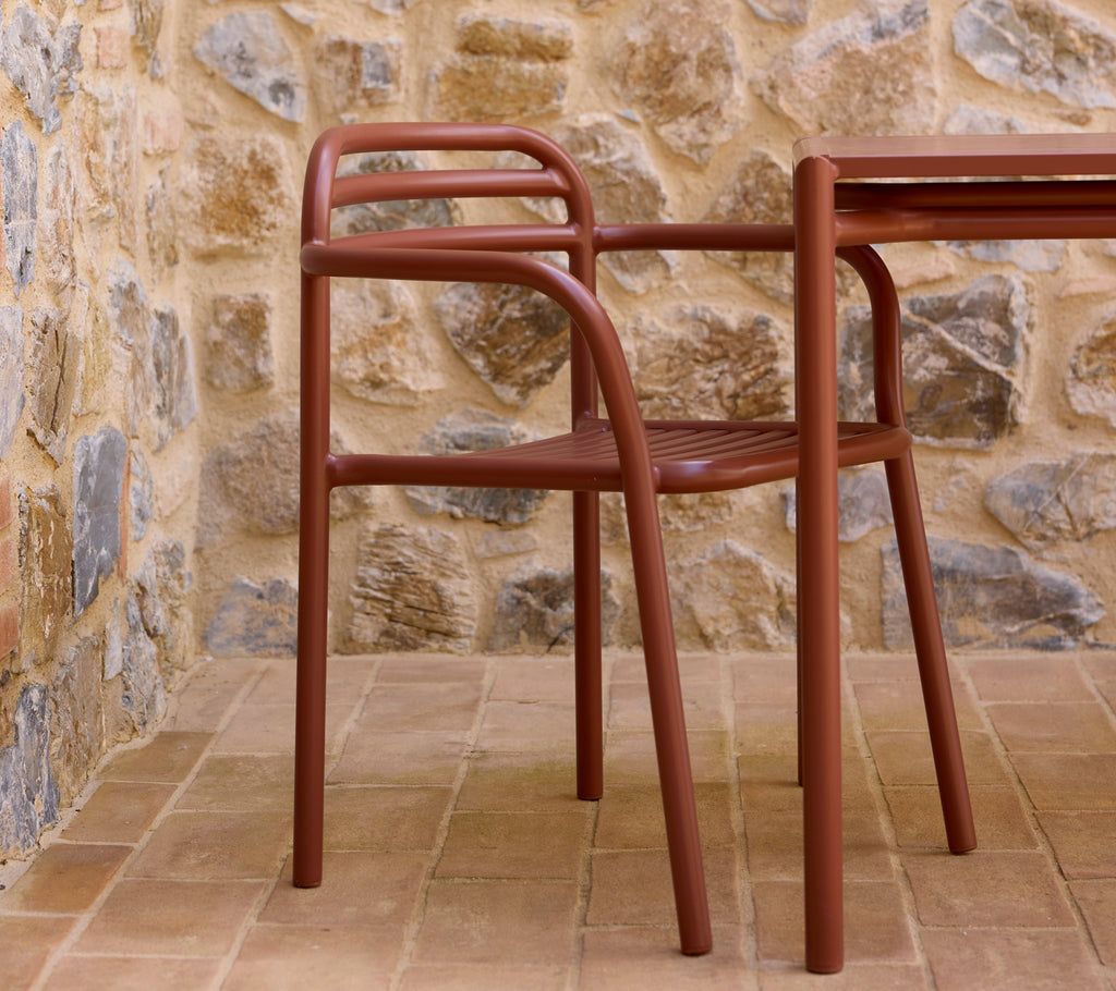 Desert red Bliss armchair next to a matching table, set against a textured stone wall. A stylish and modern seating arrangement.
