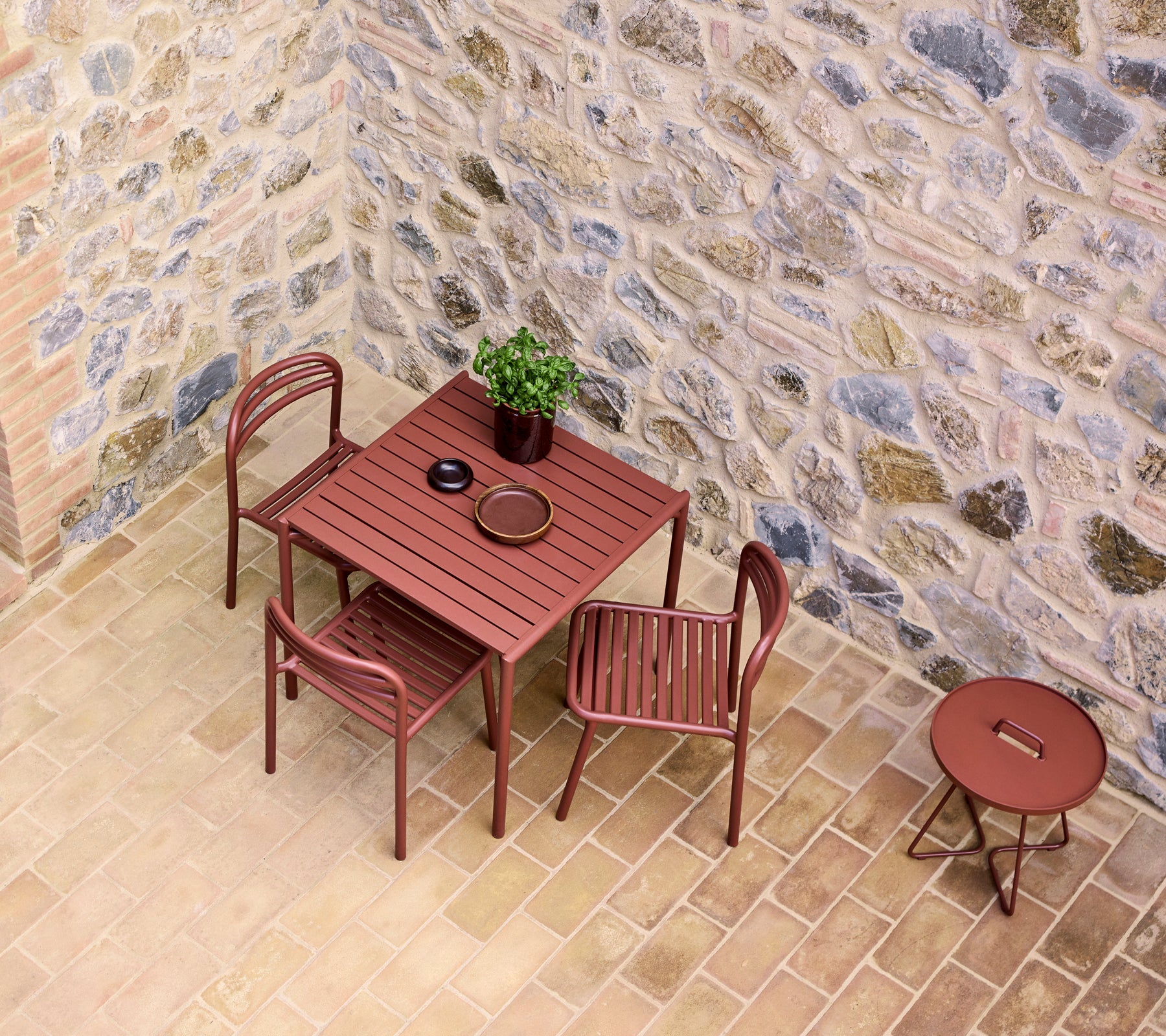 Table and chairs in a desert red hue, alongside a small round table, set against a textured stone wall with a potted plant.
