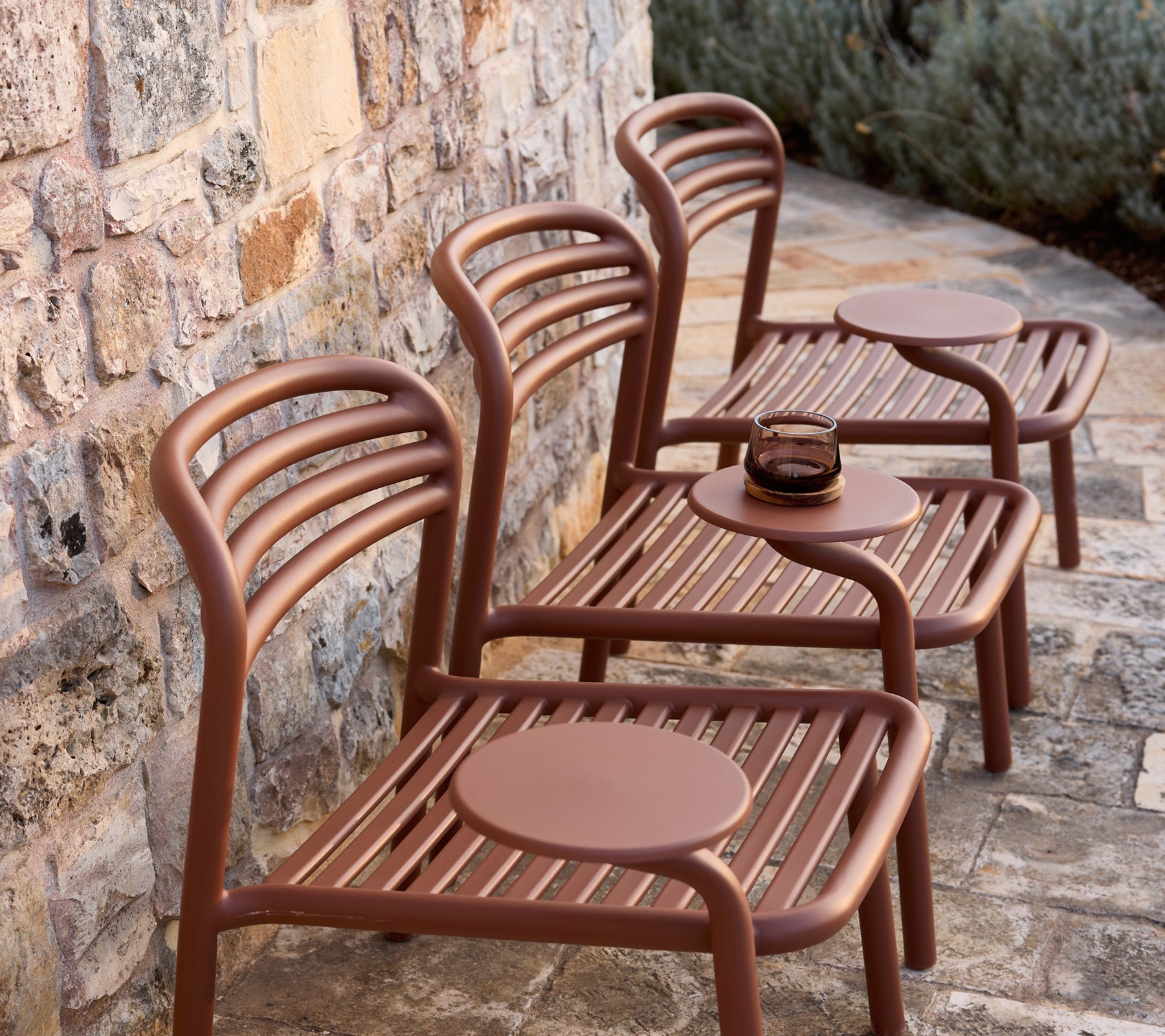 Three modern brown chairs with round tables against a stone wall.