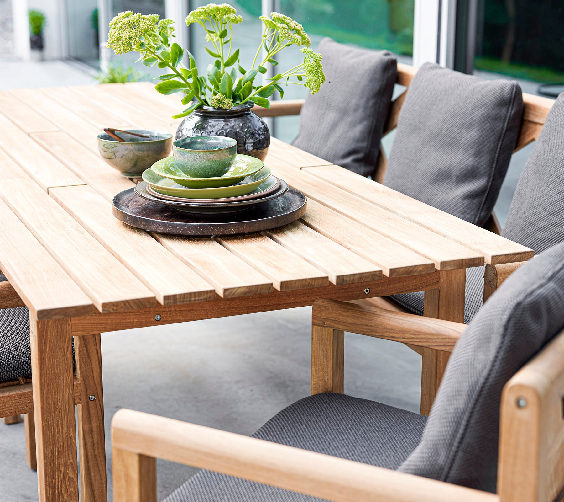 Stylish wooden table set with cushions, plates, and bowls, accented by a vase with greenery. Perfect for inviting gatherings.