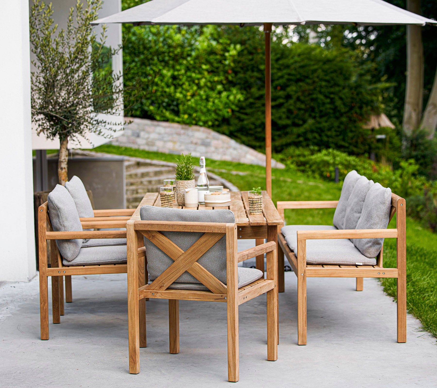 Wooden dining set featuring a rectangular table and upholstered chairs, with a large umbrella providing shade. Greenery is visible in the background.