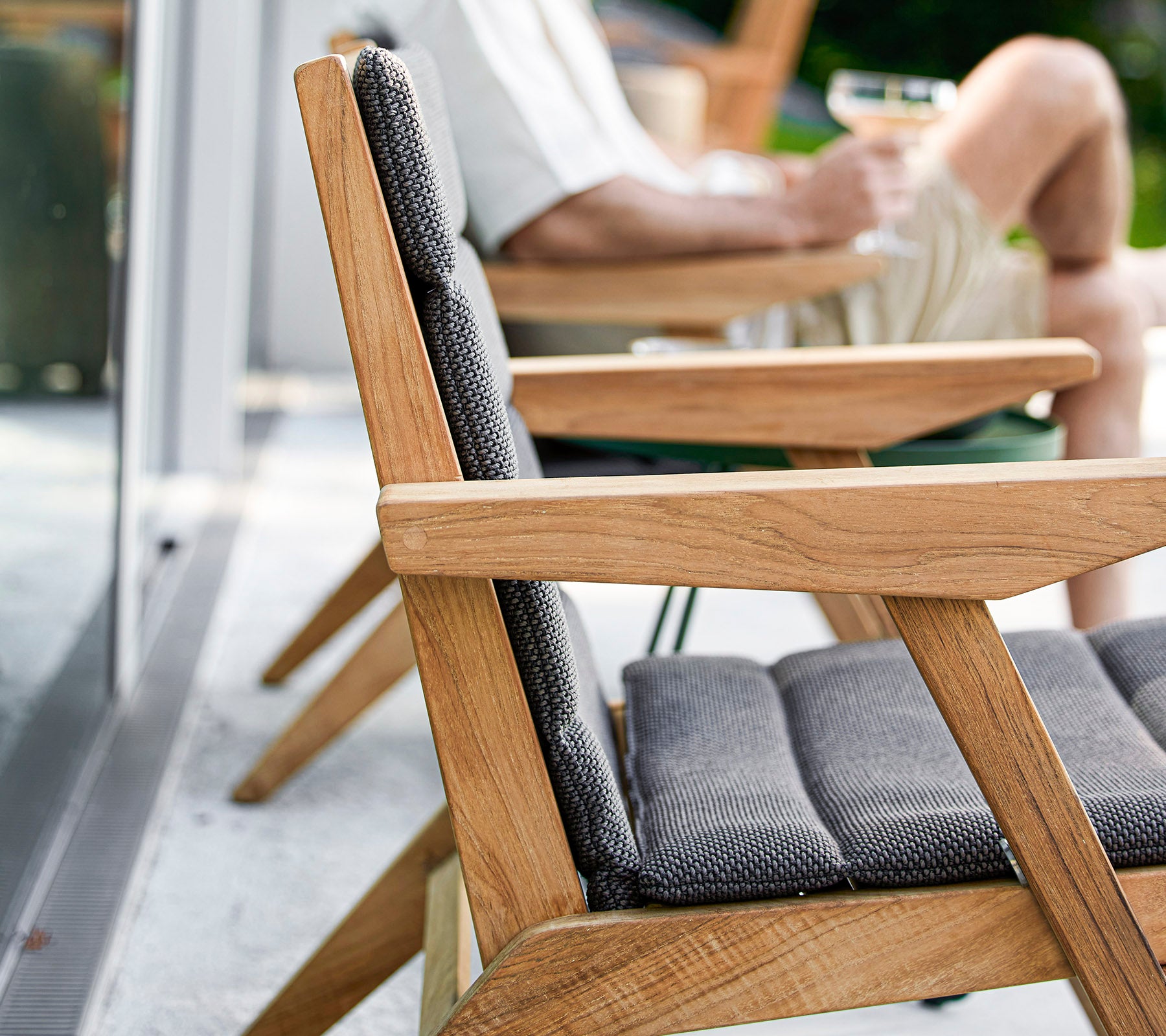 A wooden chair with a grey cushion in focus, with a person relaxing nearby holding a drink, capturing a moment of leisure.
