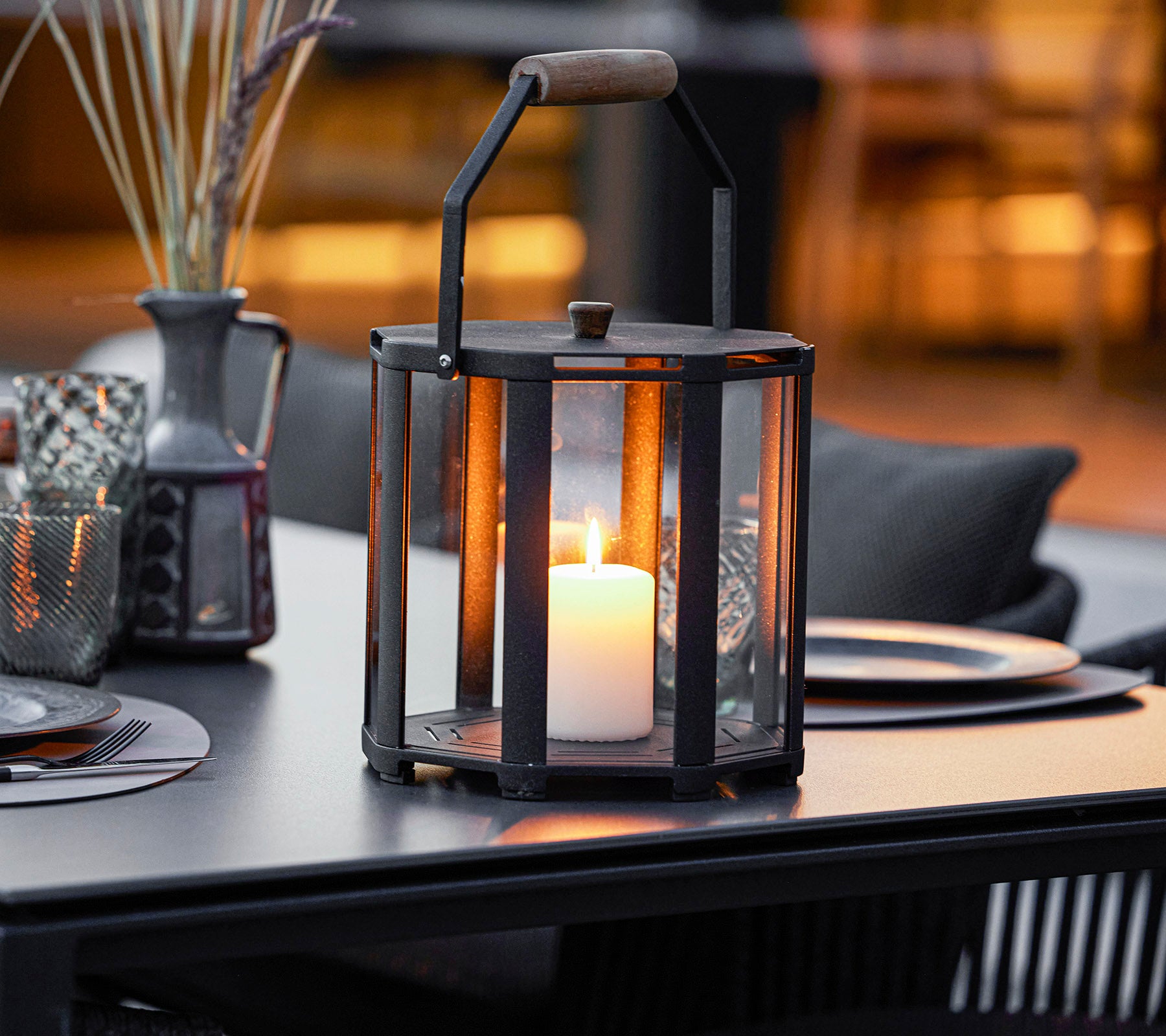 A stylish lantern with a candle inside, featuring a metal frame and wooden handle, placed on a table with decorative elements around it.