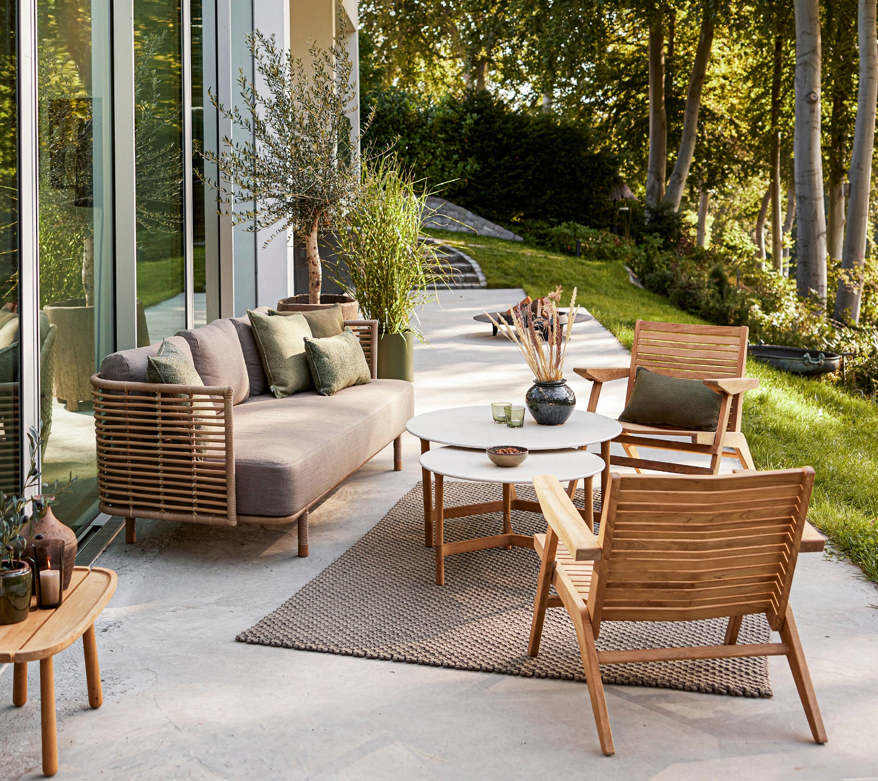 Stylish outdoor seating area featuring a modern sofa, wooden chairs, a round table, and decorative plants set against a serene backdrop.
