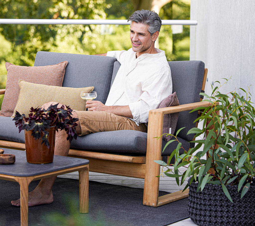 A relaxed man sits on a stylish sofa with cushions, enjoying a drink beside a potted plant and a coffee table.