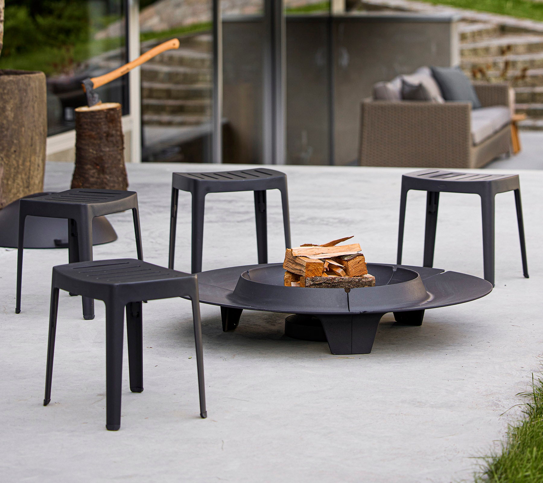A circular fire pit surrounded by four sleek black stools, with a wood stack placed in the center. Modern outdoor gathering space.