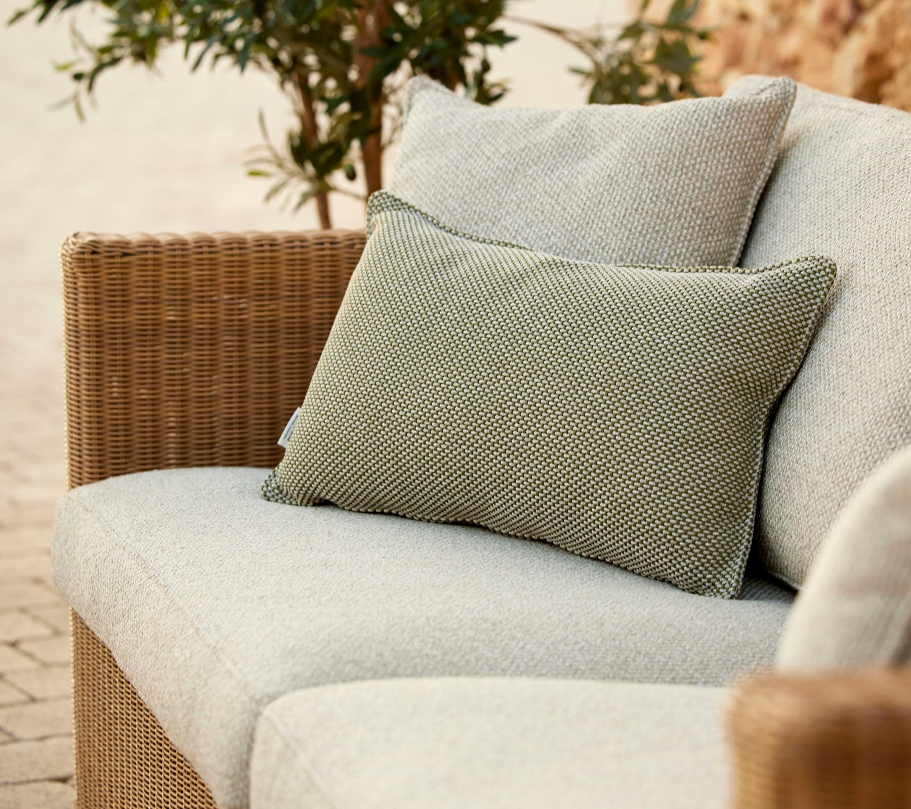 A stylish sofa featuring a light gray cushion and a textured green pillow, surrounded by greenery. Perfect for relaxation.