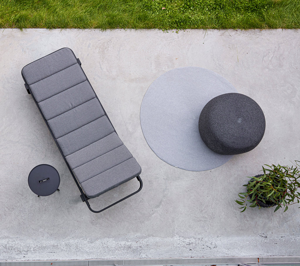 A circular footstool and a cut sunbed are arranged beside a small table, with greenery in the background.