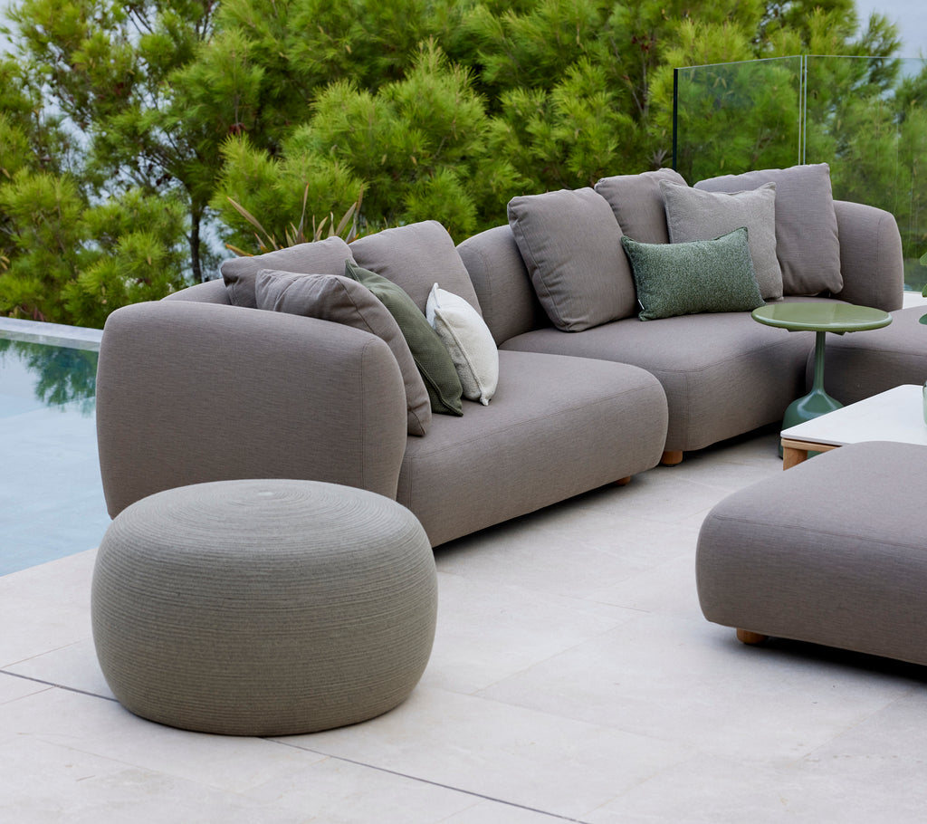Large taupe circle footstool next to a modern sofa with green cushions, surrounded by lush greenery and a serene atmosphere.