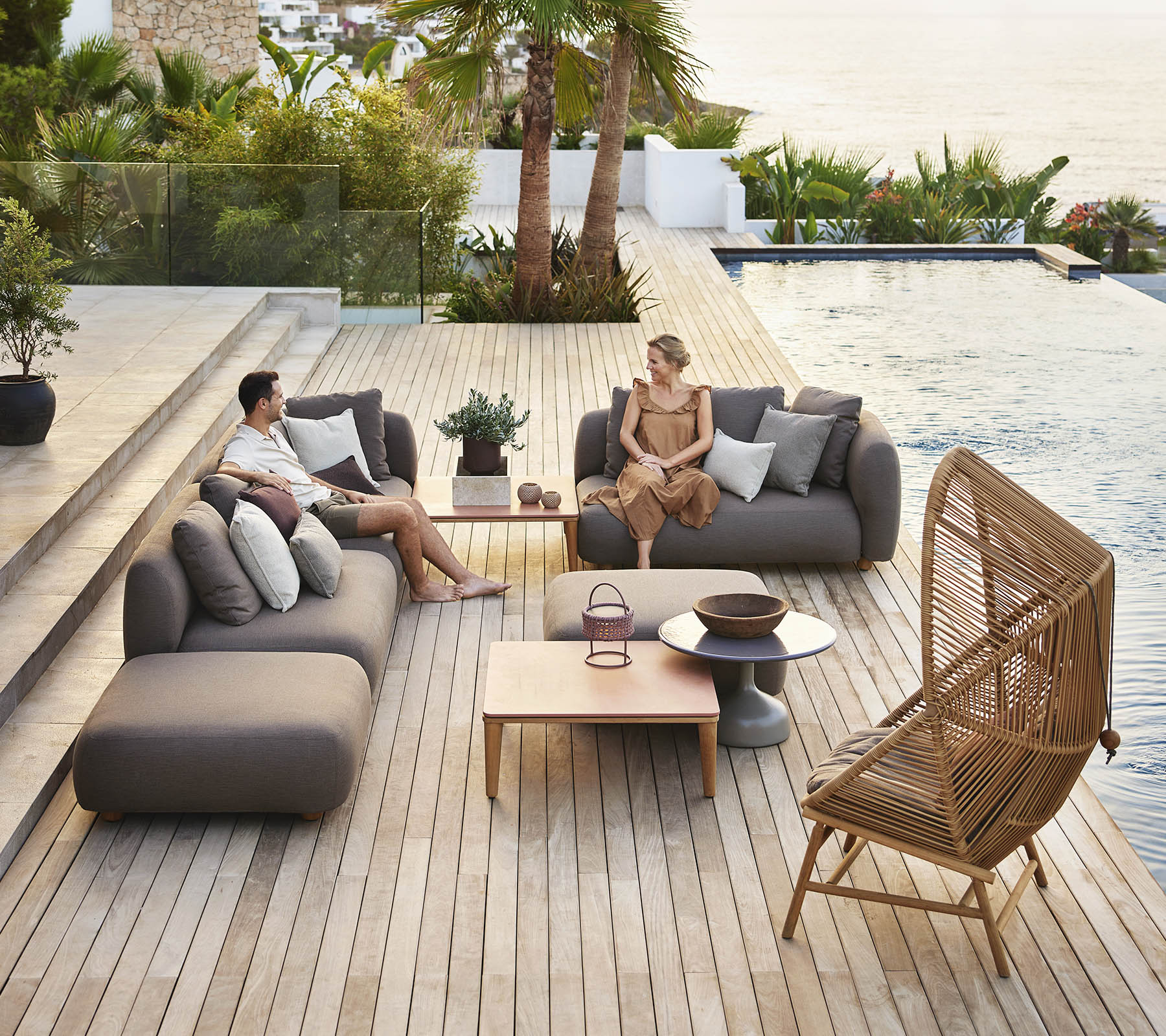 Two people relax on modern seating by a pool, surrounded by greenery, with a wooden table and decorative plants nearby.