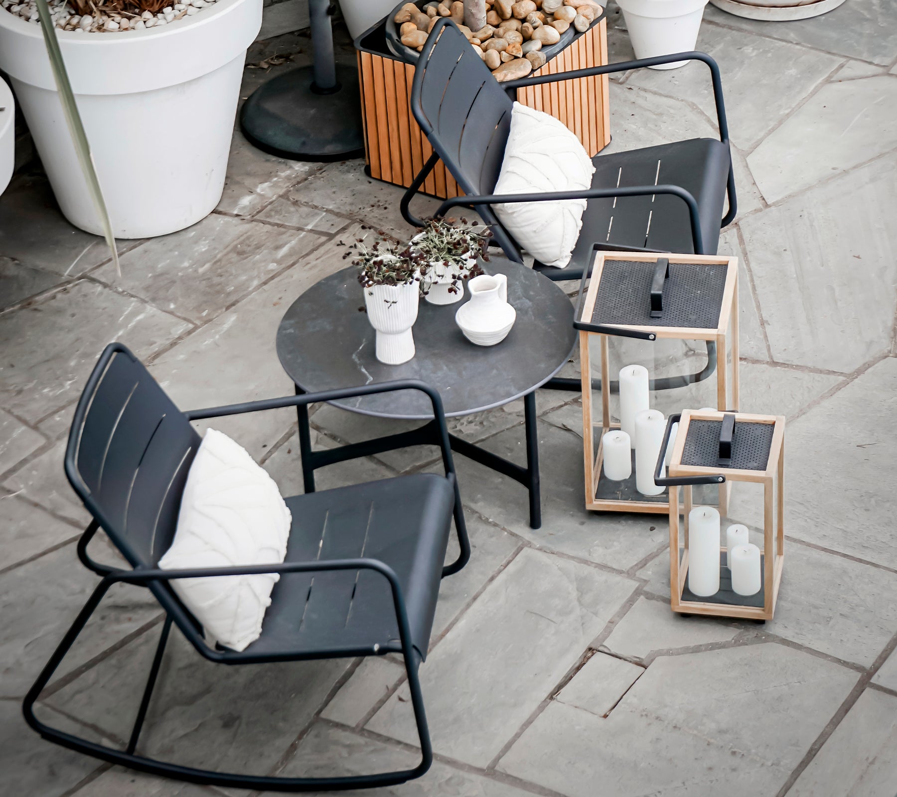 Two modern rocking chairs and a round table with decorative plants and lanterns, set on a textured stone surface.