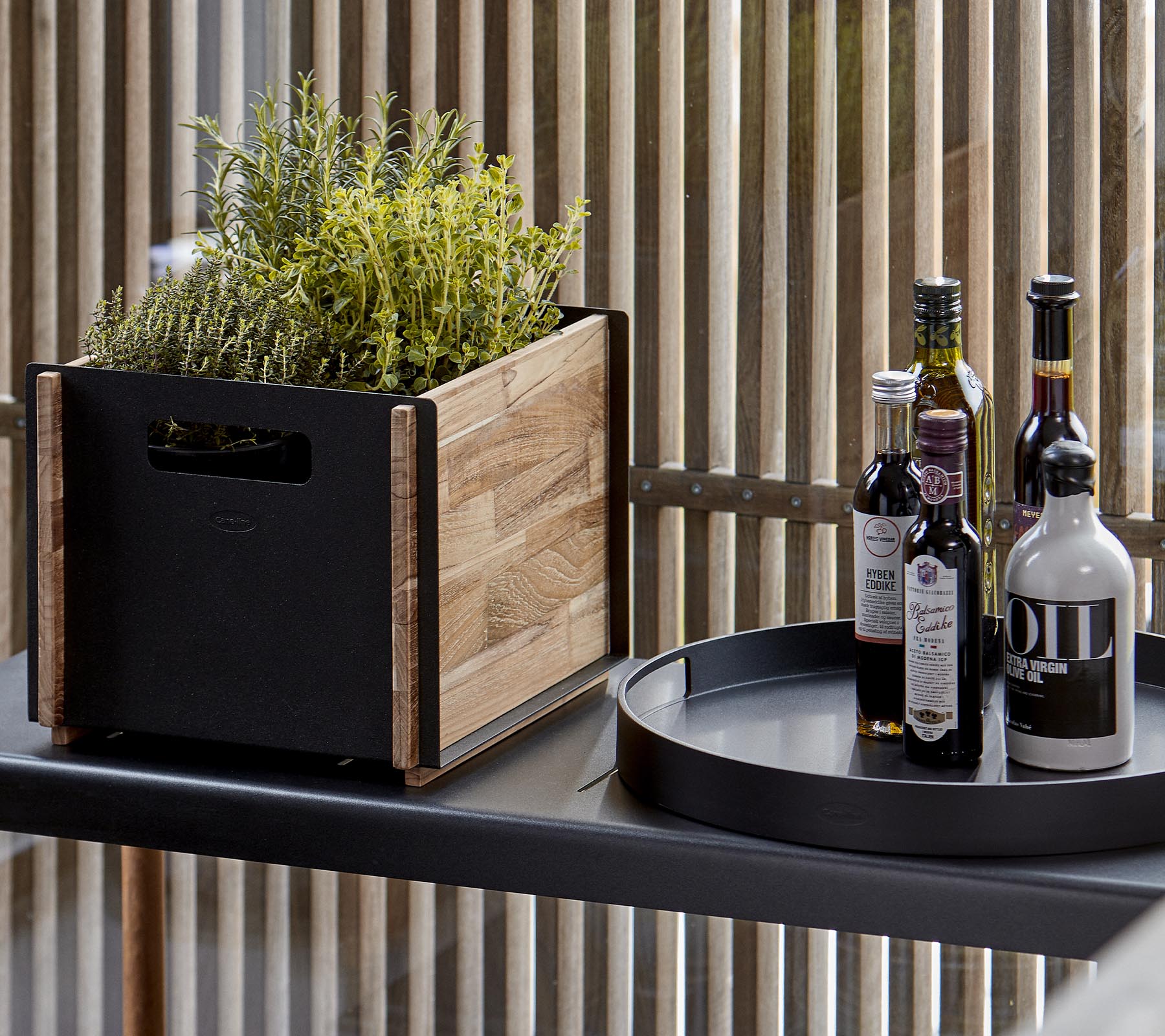 A wooden planter with greenery beside a black circular tray holding several bottles of oil and vinegar.